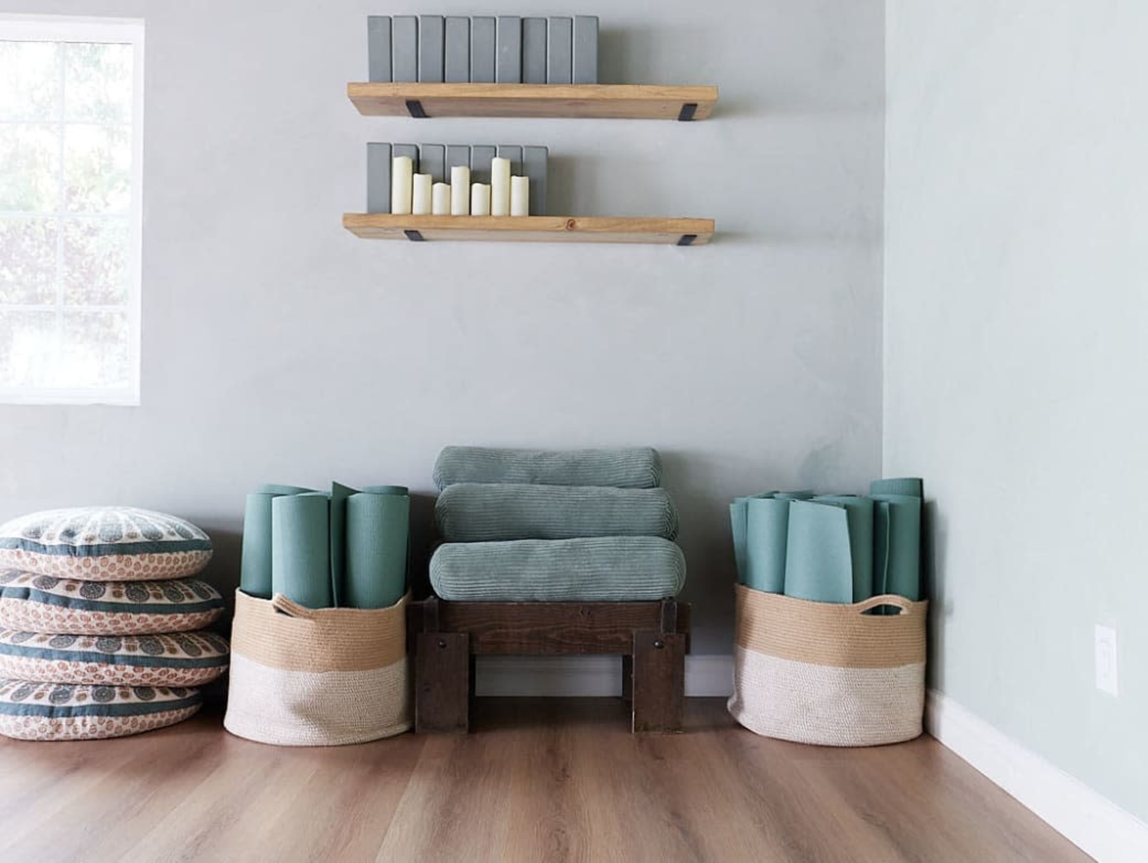 The image shows a minimalist room with yoga mats stored in baskets, a wooden bench, and shelves displaying candles and books.