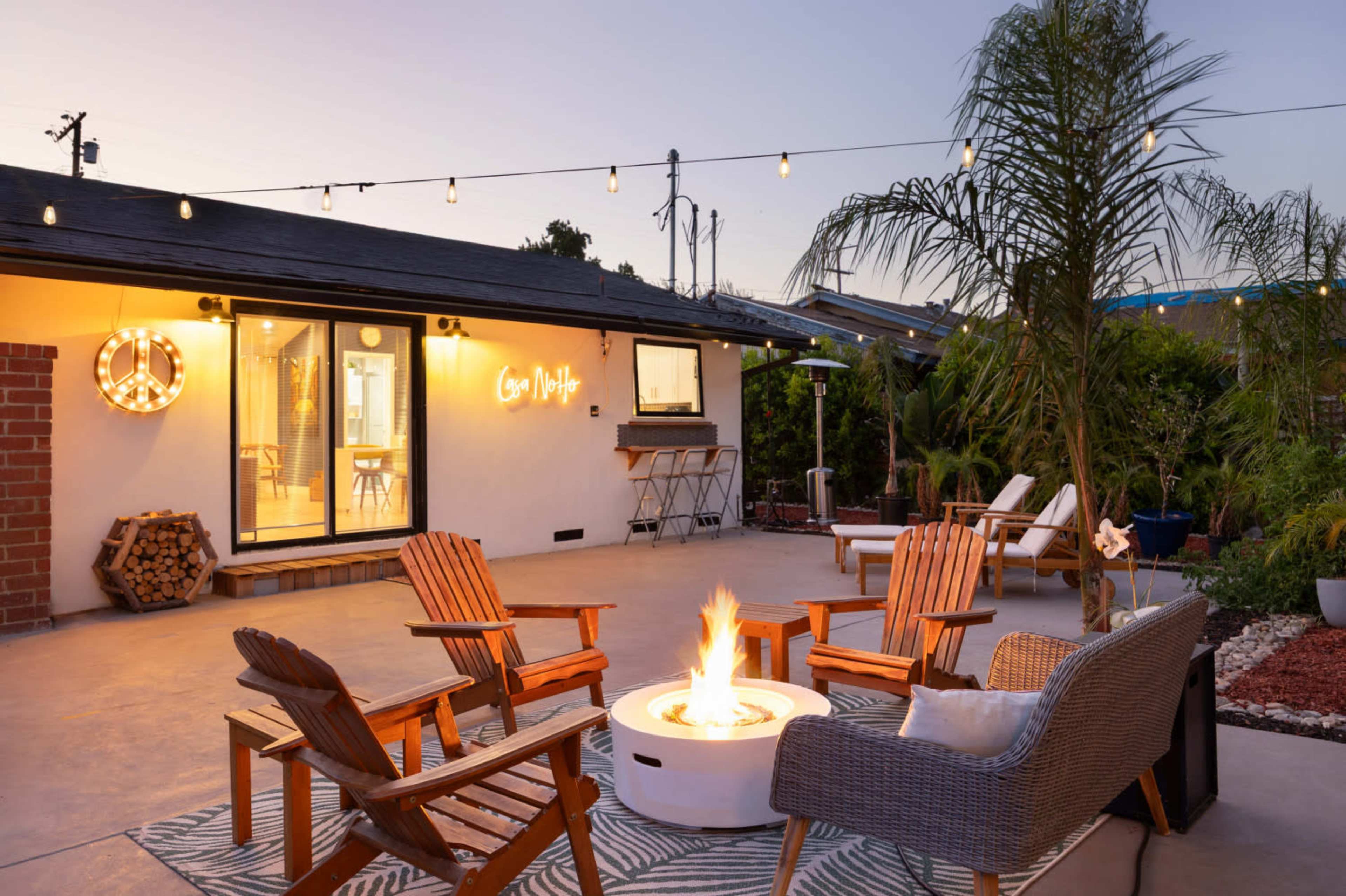 A modern backyard features a fire pit surrounded by wooden chairs, against a backdrop of string lights and a well-lit house with patio furniture.
