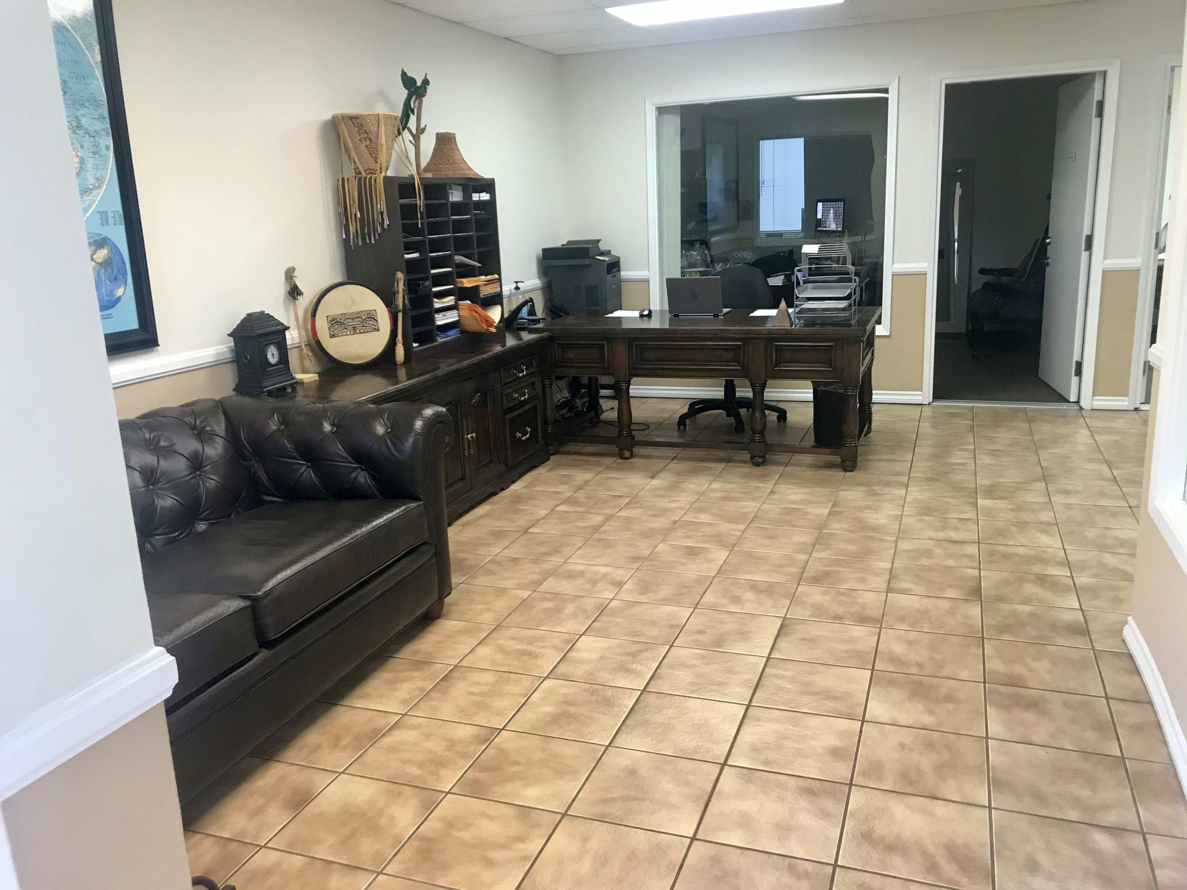 An office interior with a leather sofa, a wooden desk with office equipment, and a glass-walled room in the background.