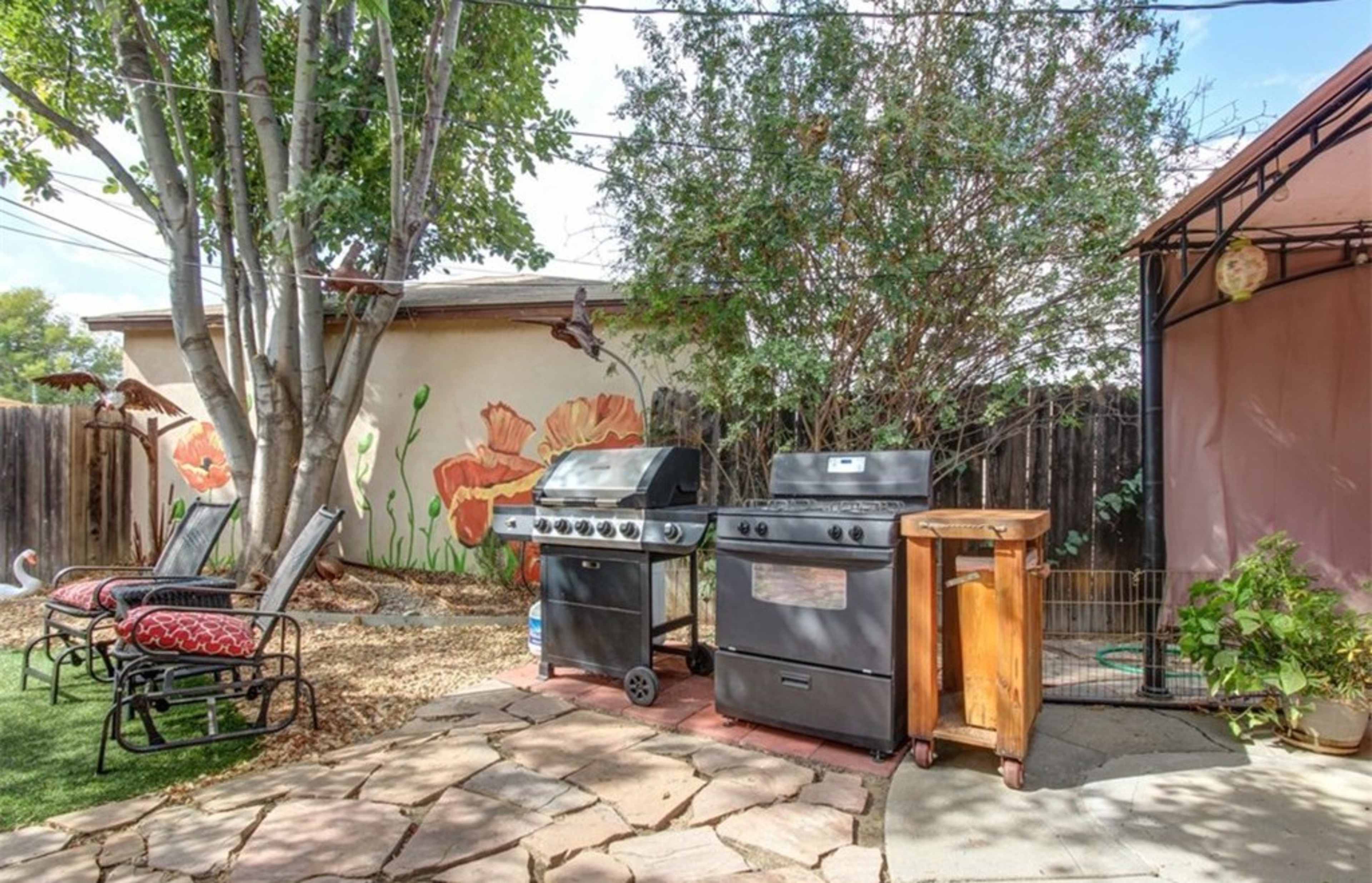 The image shows a small outdoor patio area featuring two grills, a wooden cart, and seating arranged on stone flooring, surrounded by greenery and a mural of flowers on the wall.