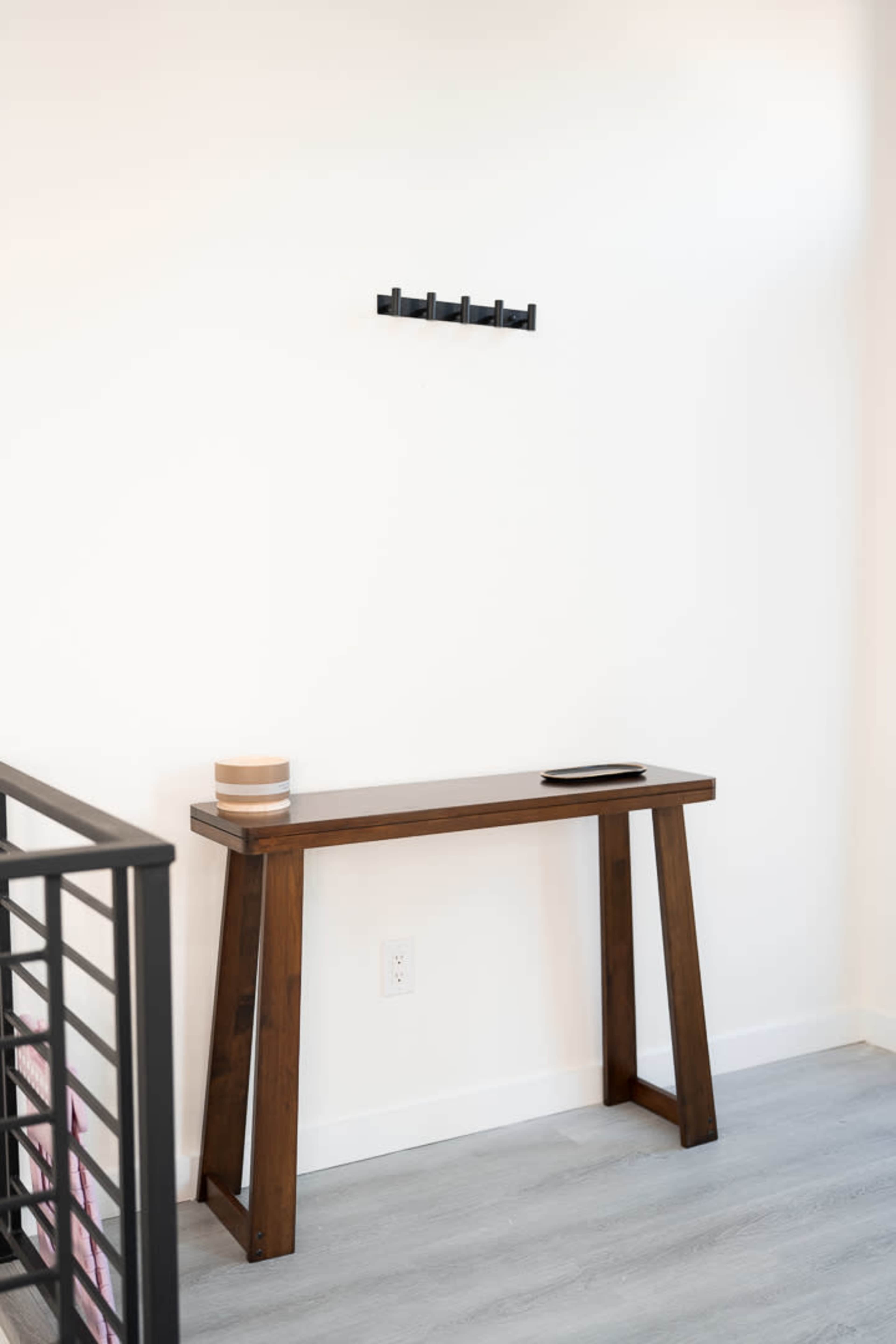 A wooden console table with two legs stands against a white wall, above which is a black wall-mounted coat rack.
