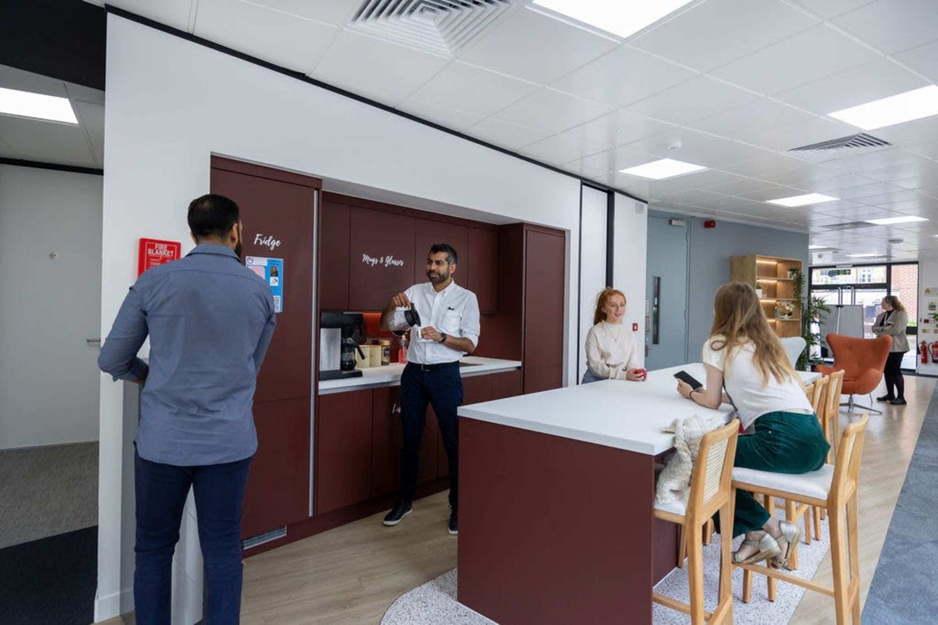 A modern office break room features a coffee station with several people engaging in conversation and using their devices.