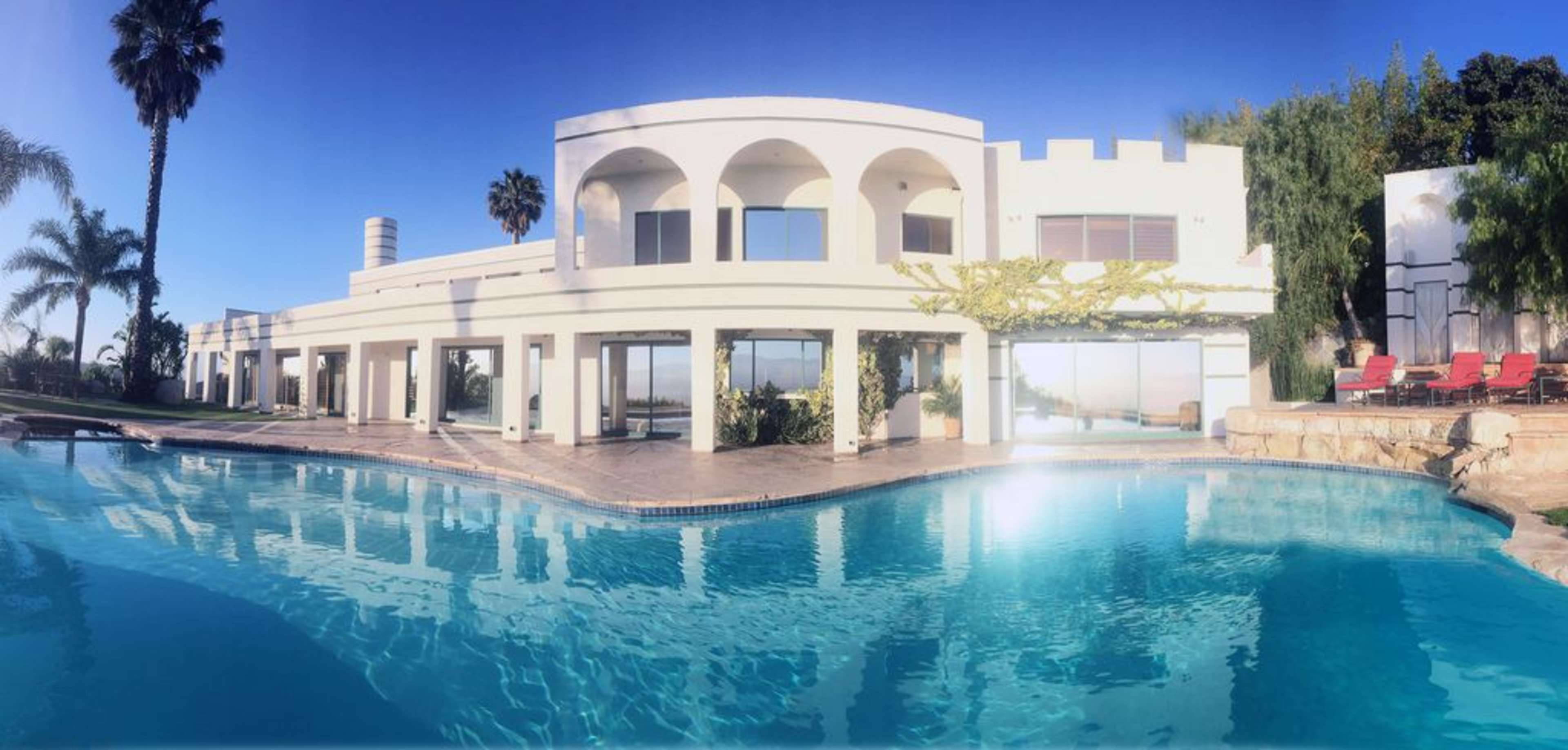 A white modern house with arched windows overlooks a swimming pool surrounded by palm trees.