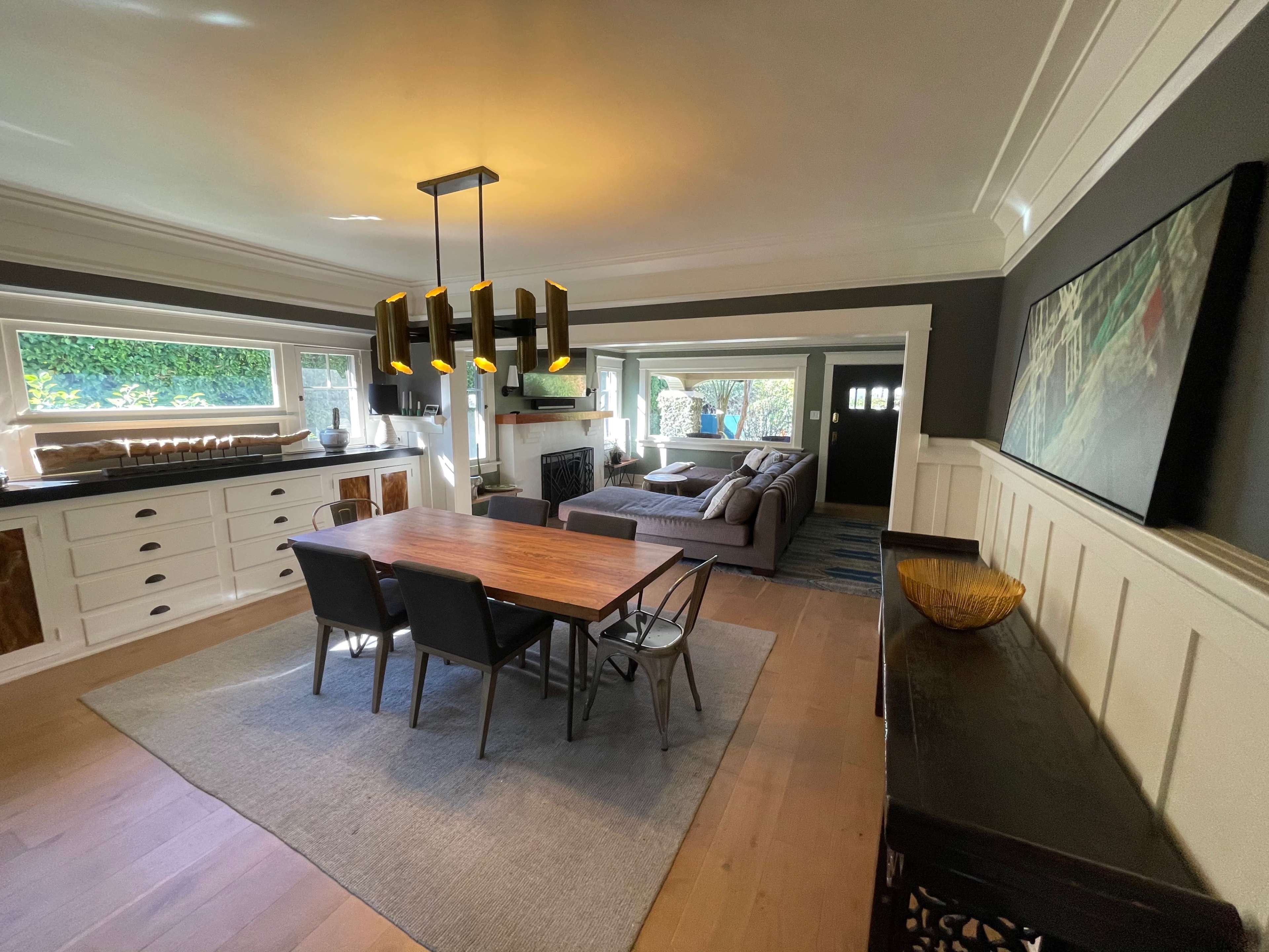 A bright living space featuring a wooden dining table with chairs, a cozy couch, large windows, and decorative elements on the walls.