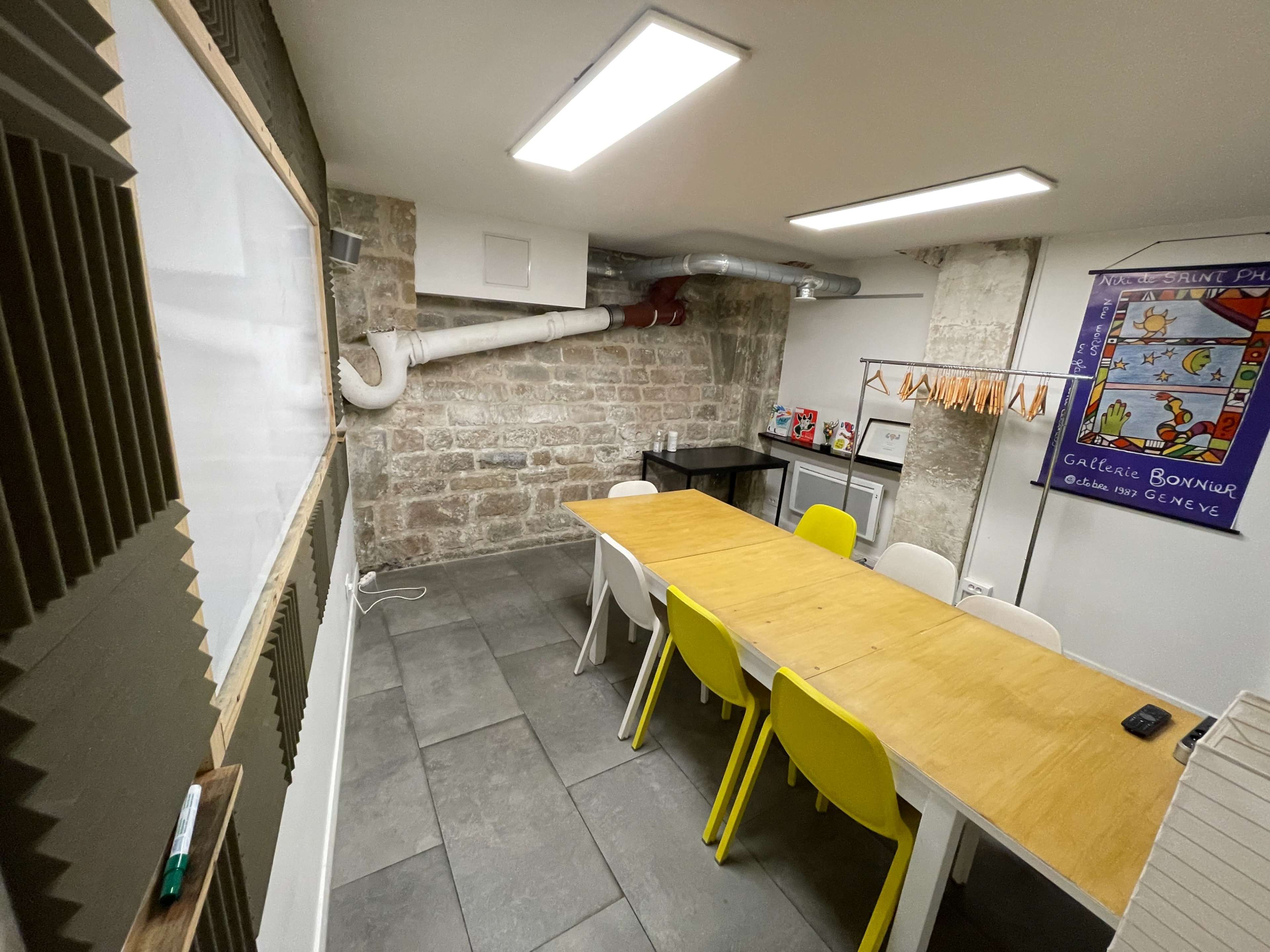 A spacious meeting room features a long wooden table surrounded by colorful chairs, with a kitchenette and whiteboard along the stone wall.