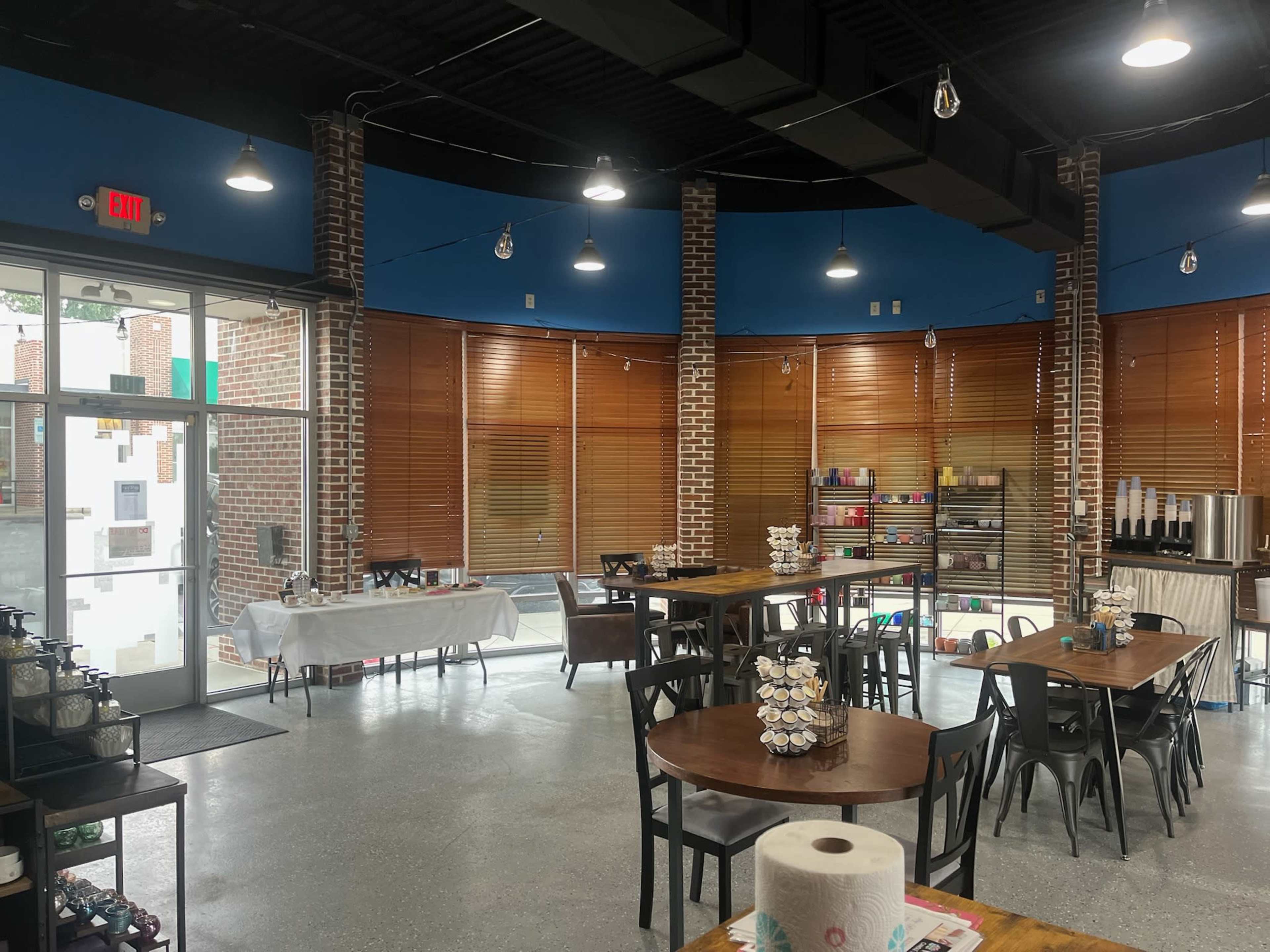 The image shows the interior of a modern café with wooden tables, chairs, and a variety of items displayed on shelves, illuminated by overhead lights and natural light from large windows.