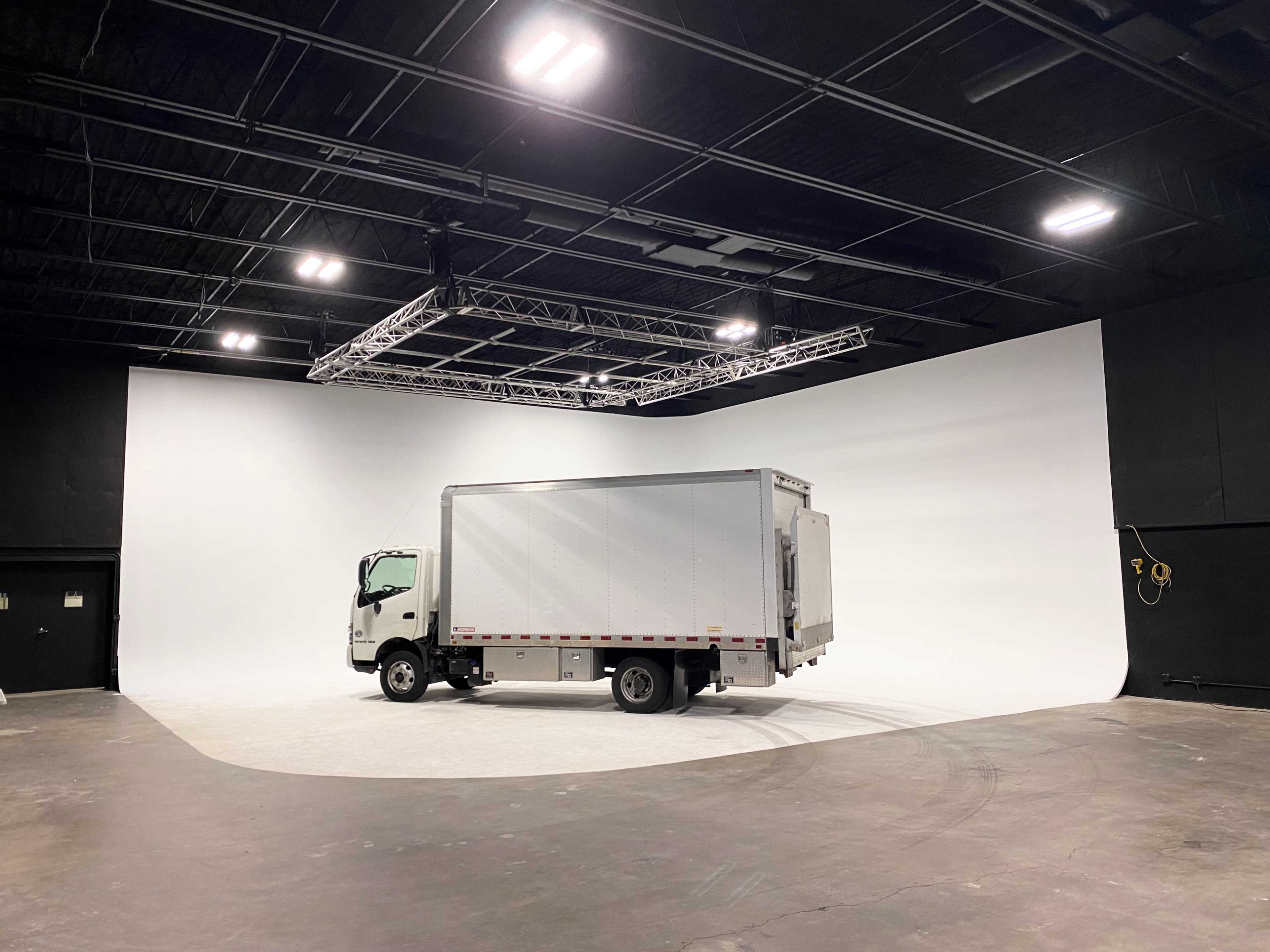 A white delivery truck is parked in a spacious studio with a curved white backdrop and industrial lighting.