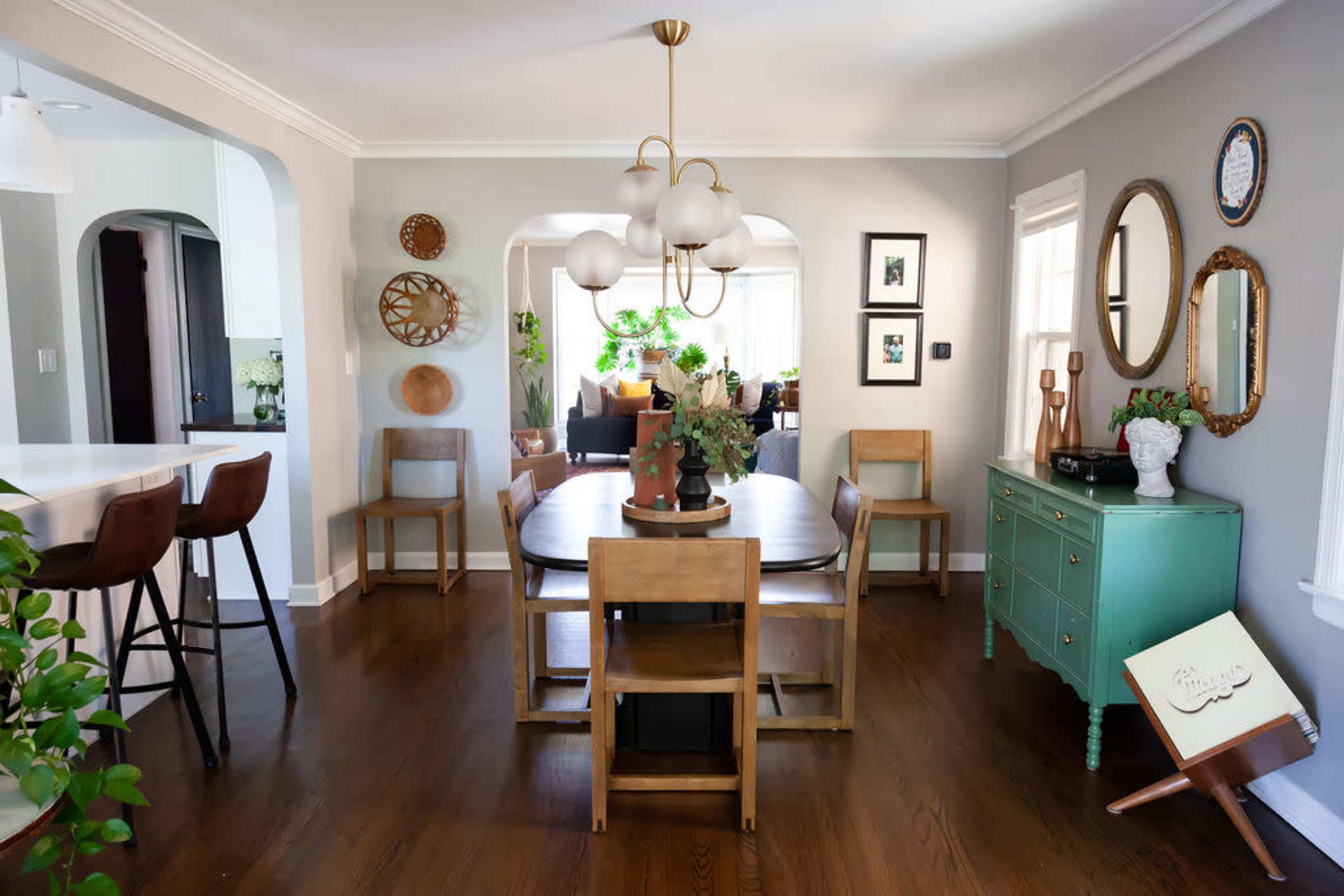 A dining area features a round table surrounded by wooden chairs, with a green sideboard and decorative wall elements such as mirrors and framed pictures.