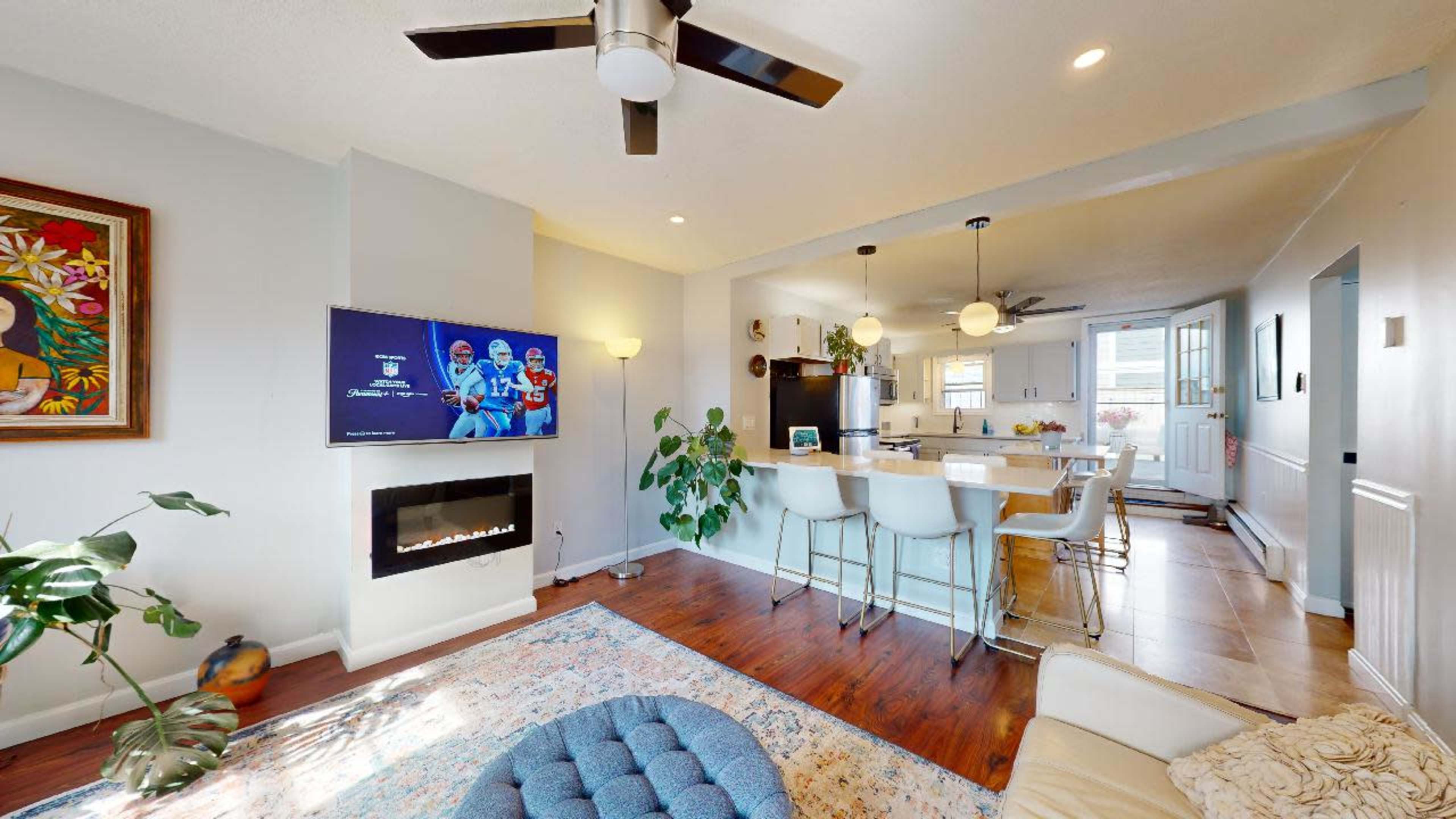 A modern living space with a wall-mounted television, a ceiling fan, a kitchen bar with stools, and a fireplace surrounded by light-colored walls and wooden flooring.