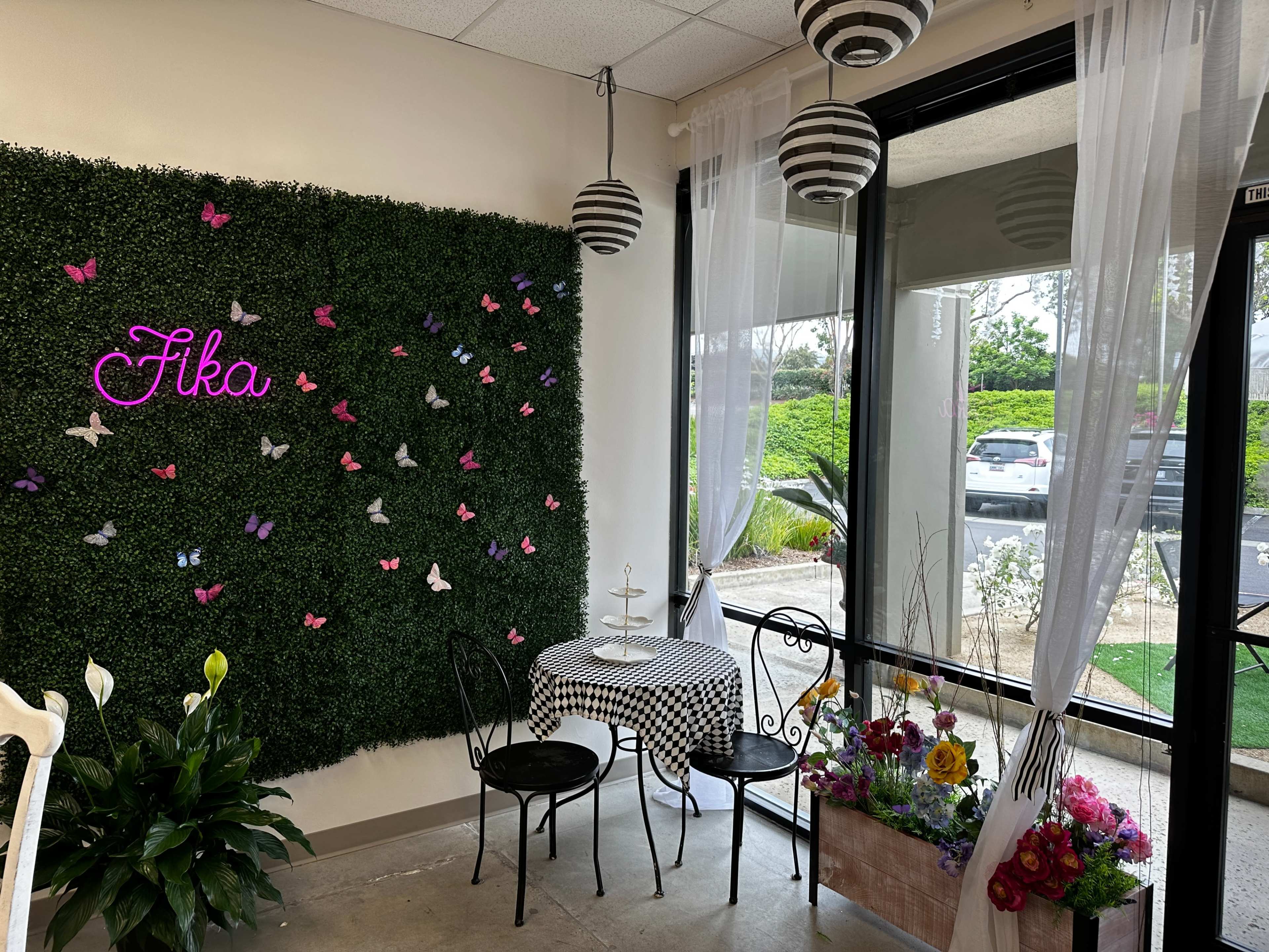 A cozy café corner with a checkered tablecloth, two chairs, a neon sign that reads "Fika," and a decorative wall covered in artificial greenery adorned with colorful butterflies.