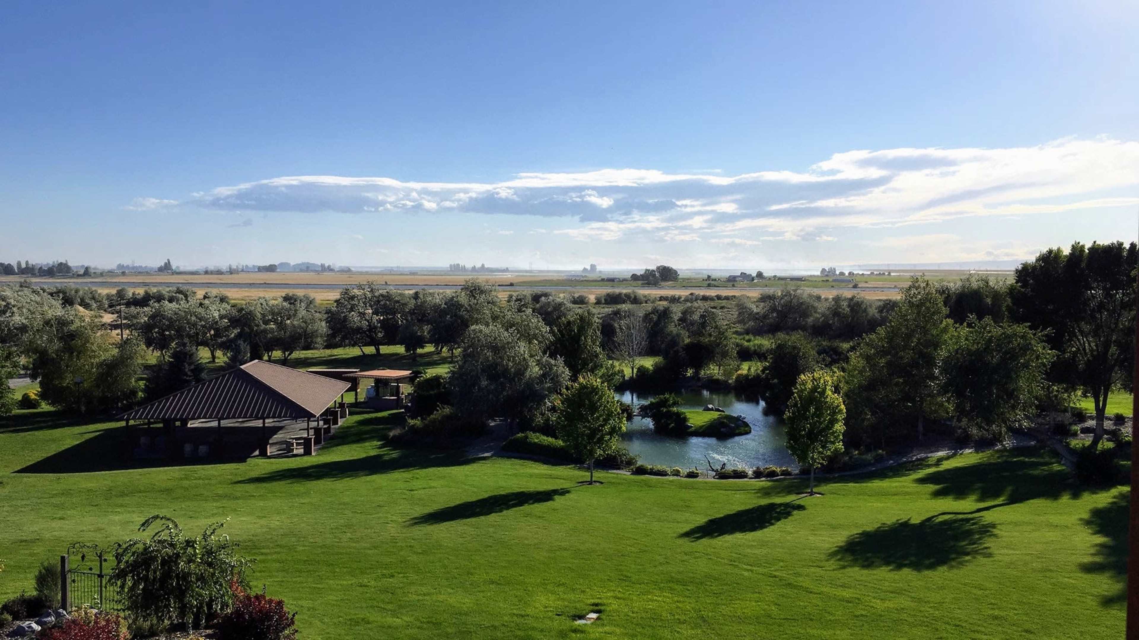 The image shows a landscaped area with a pond, trees, and a gazebo, set against a wide open landscape under a clear sky.