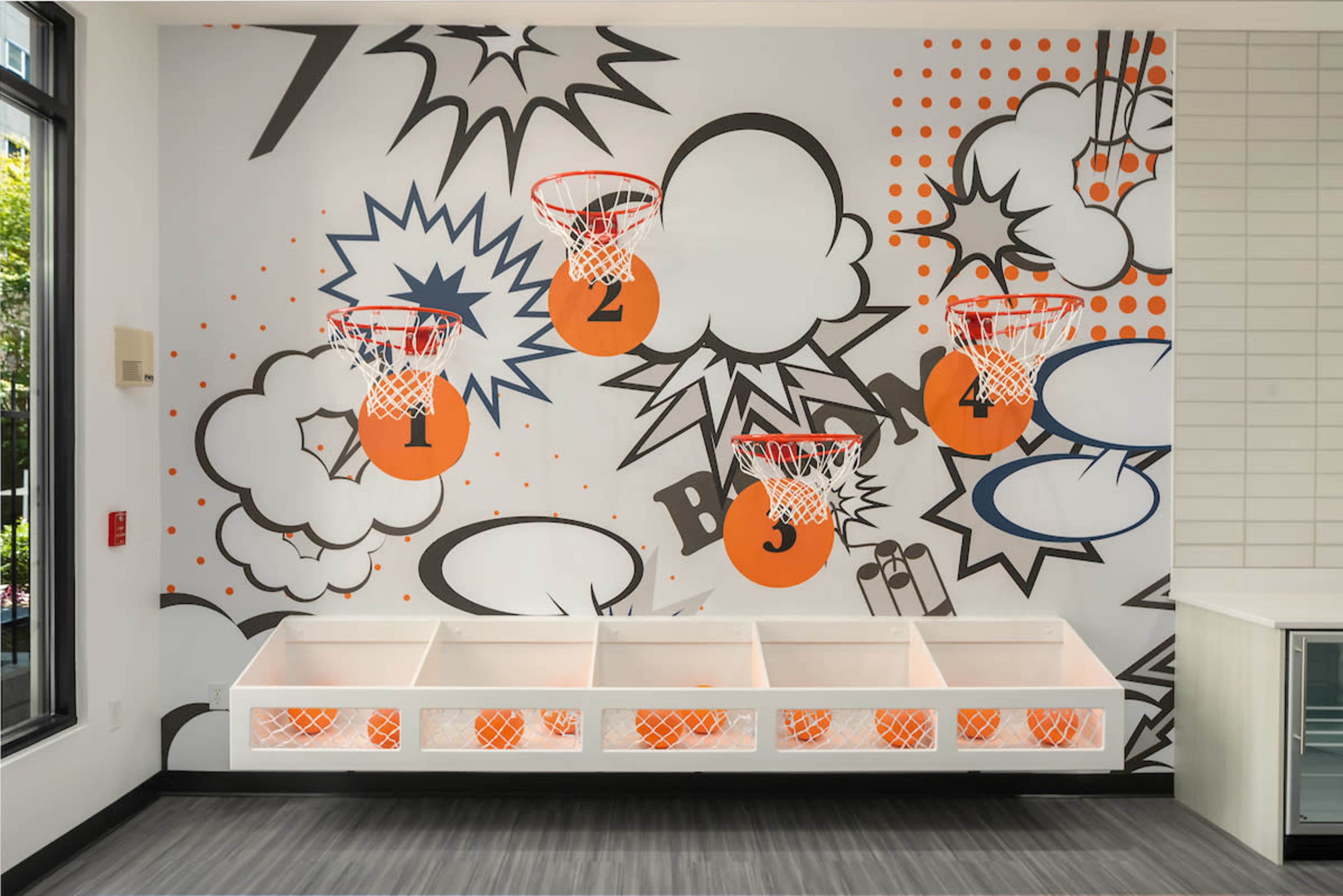 A playful interior wall featuring five basketball hoops designed with comic-style graphics and bright colors, alongside a low, white storage unit.