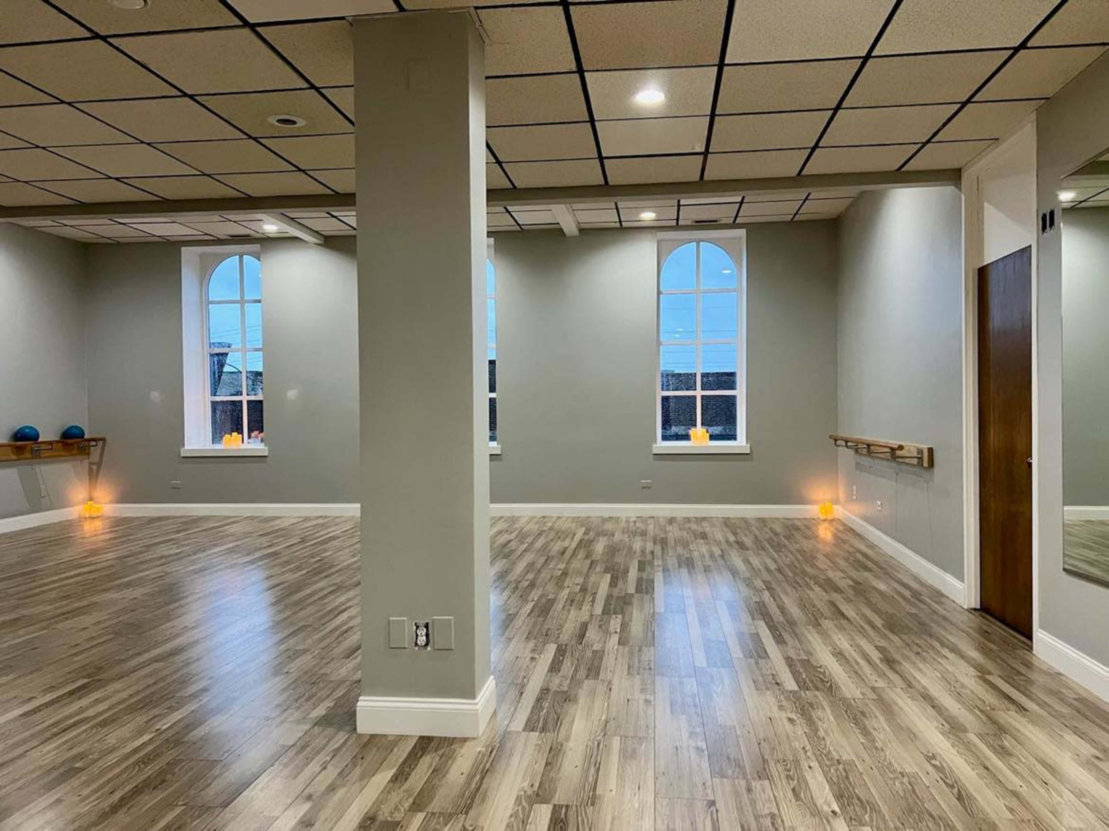 A spacious, empty fitness studio with wooden floors, large windows, and soft lighting.