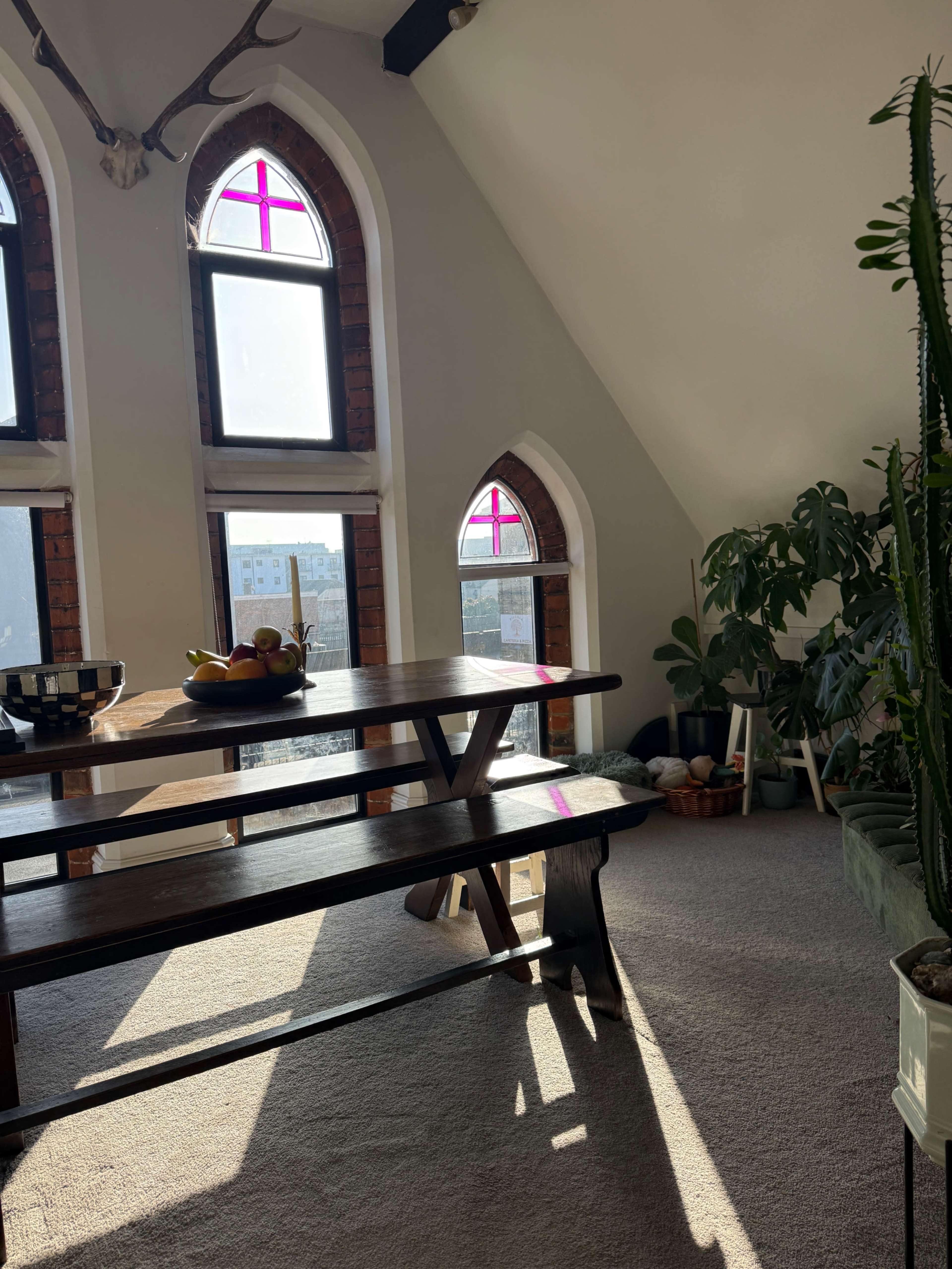 A dining area features a wooden table with a bowl of fruit, surrounded by large windows with colorful cross-shaped decorations and greenery in the corners.