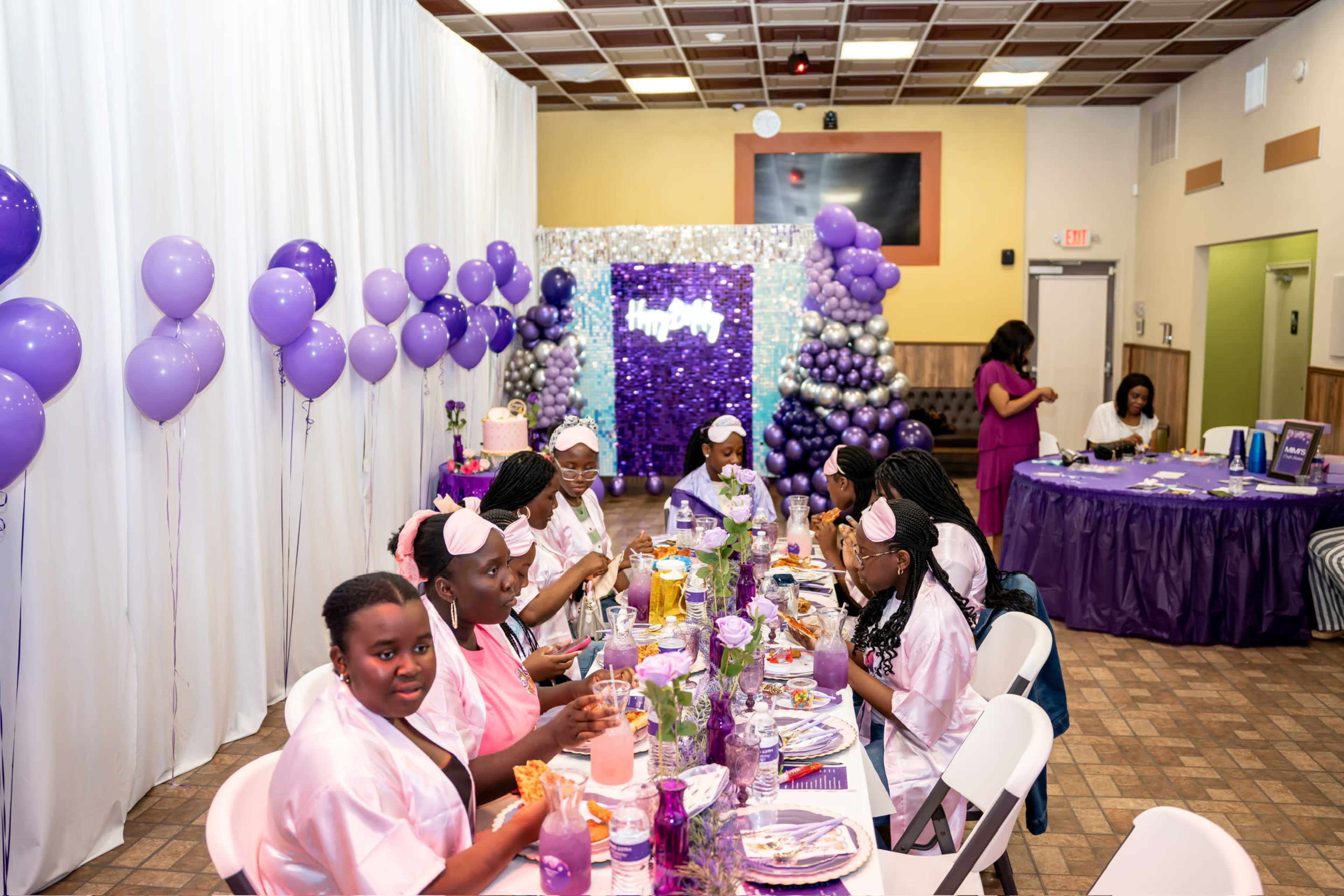 A group of young girls in matching outfits sit at a decorated table set for a party, surrounded by purple balloons and a cake display in the background.