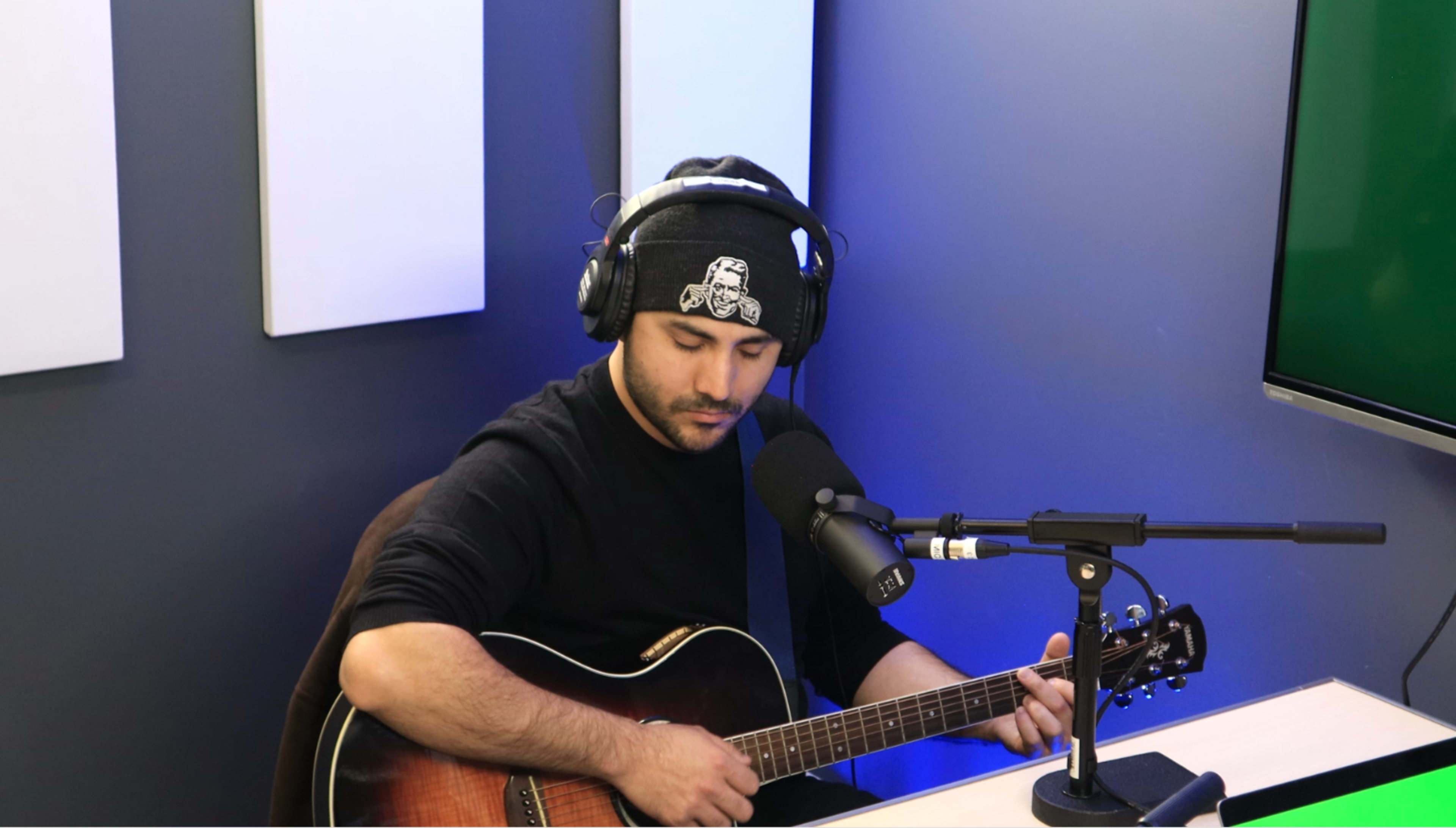 A man wearing headphones plays an acoustic guitar while seated at a table in a studio setting.
