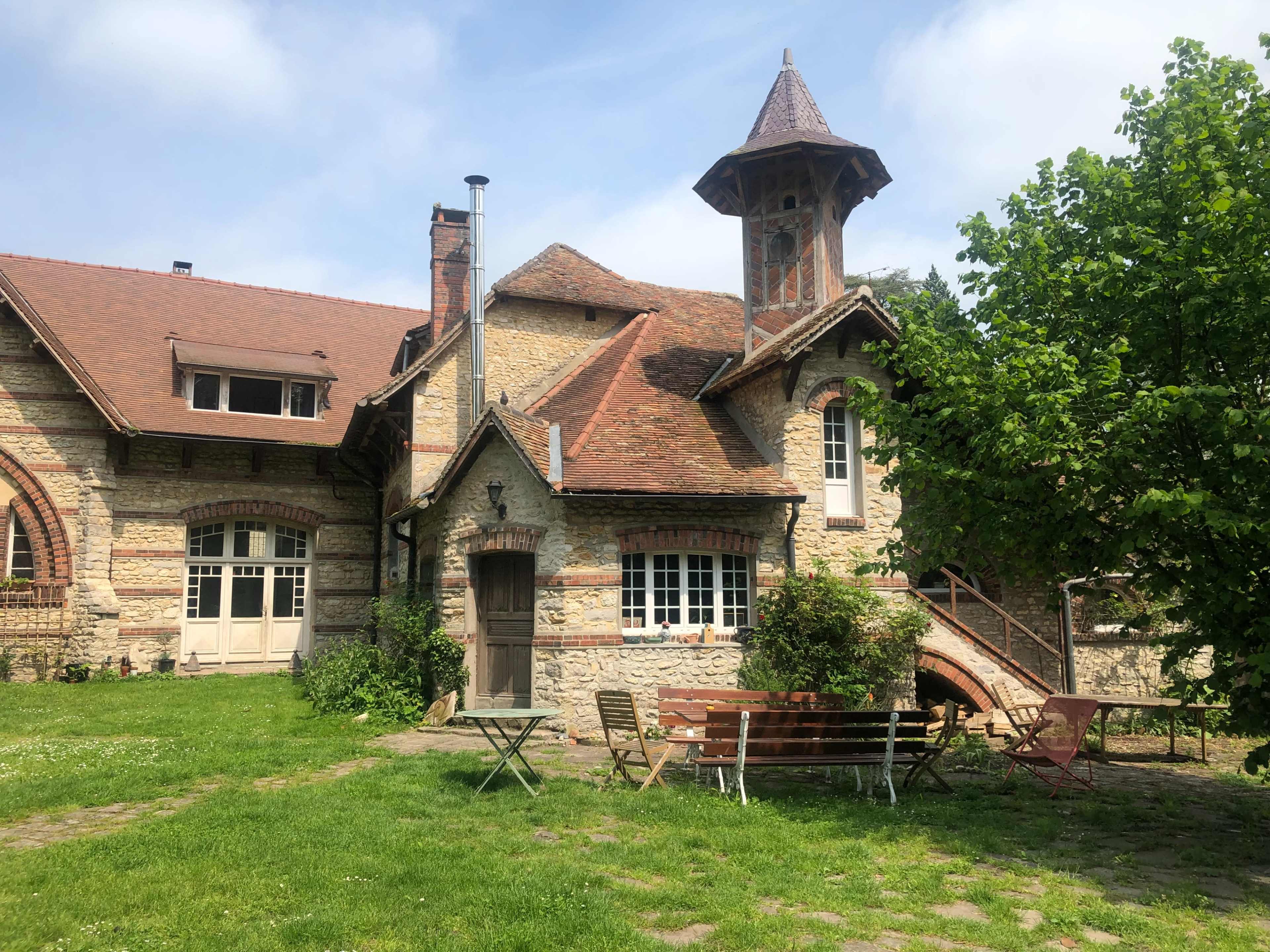 A stone house with a tower features a sloped roof and is surrounded by a garden with seating.