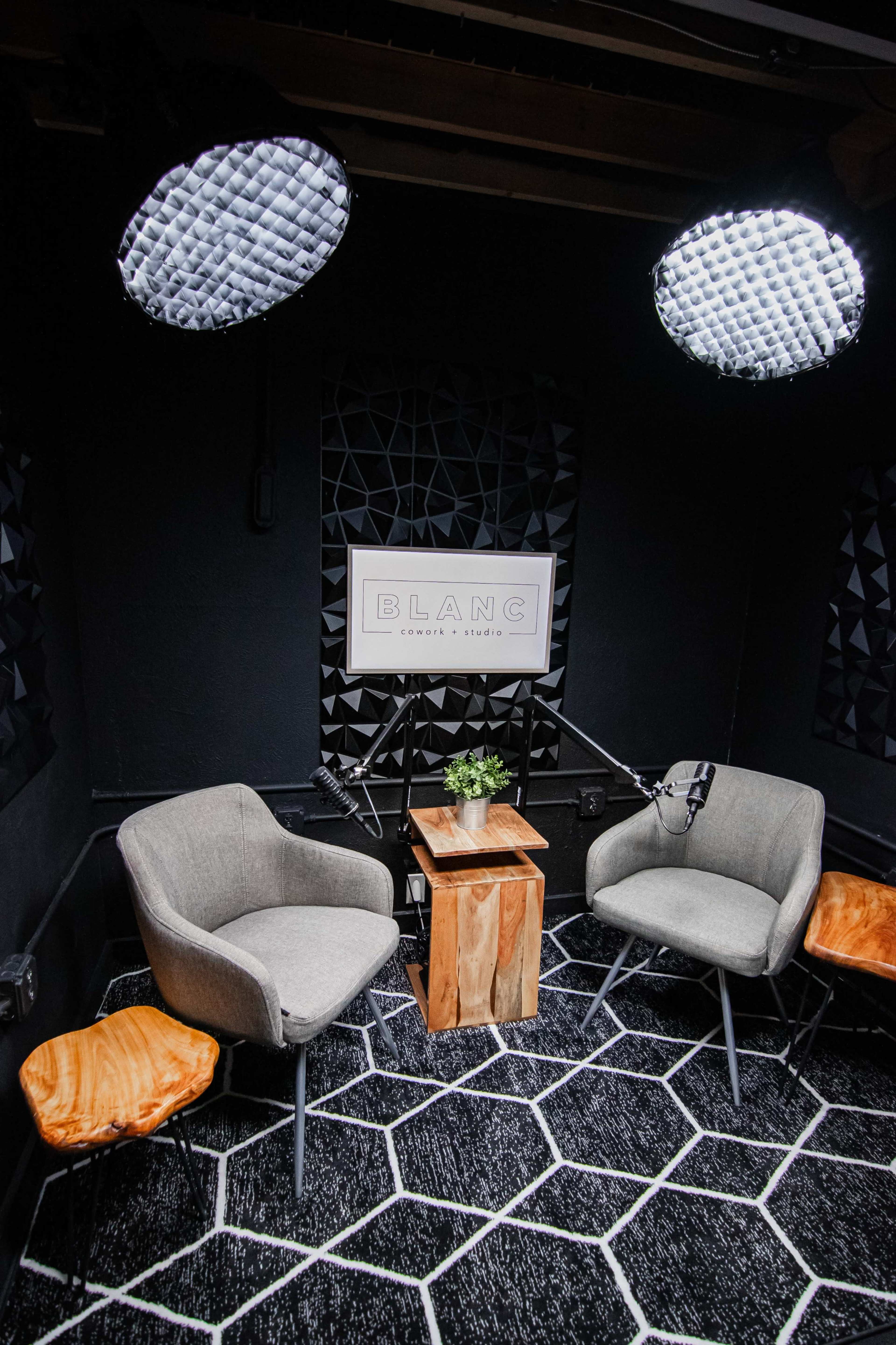 The image shows a small recording studio with two gray chairs, a wooden table with a plant, and two overhead lights.