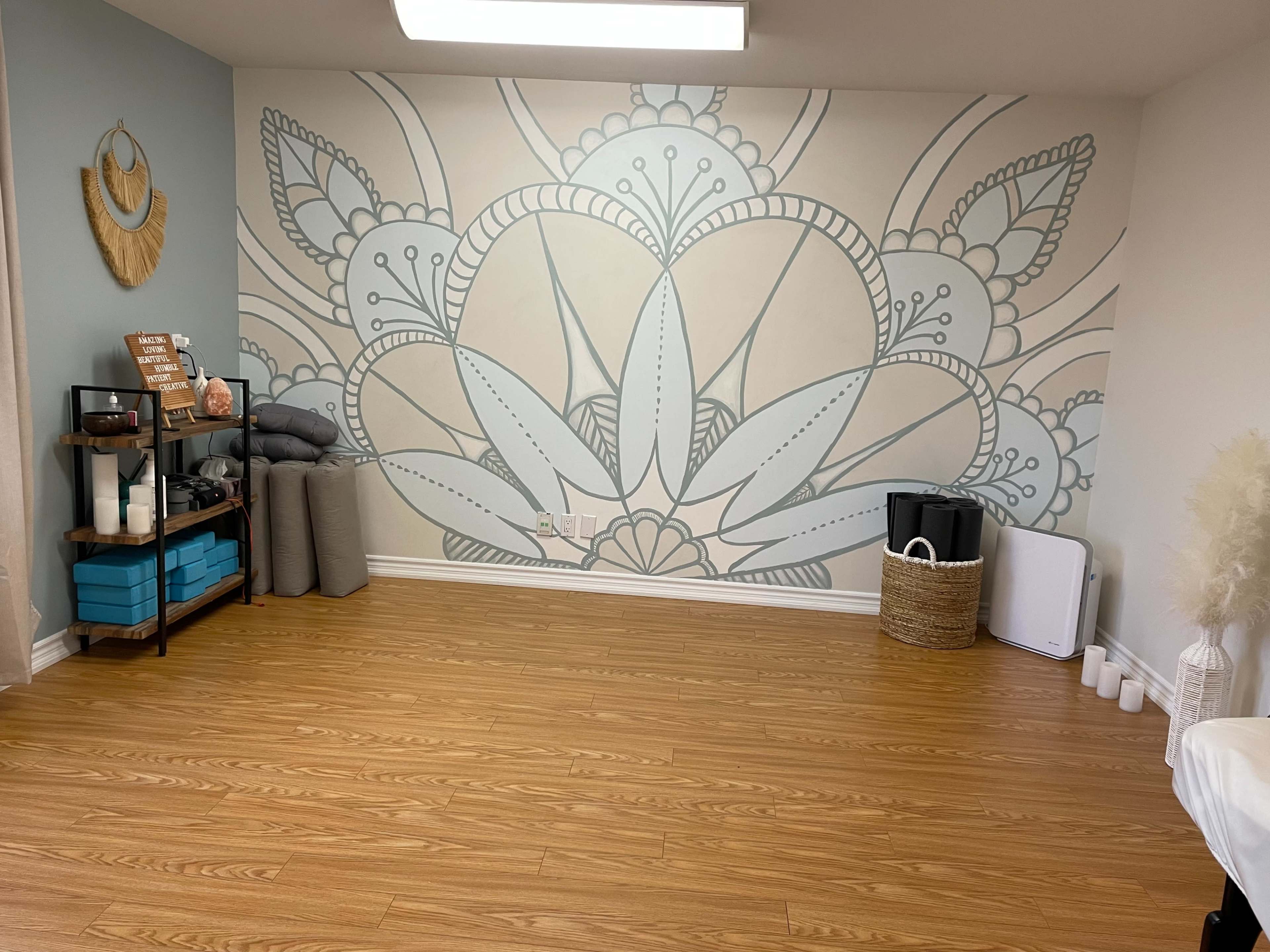 The image shows a spacious, minimalist room featuring a large geometric mural on one wall, with a wooden shelf displaying decor items and storage containers.