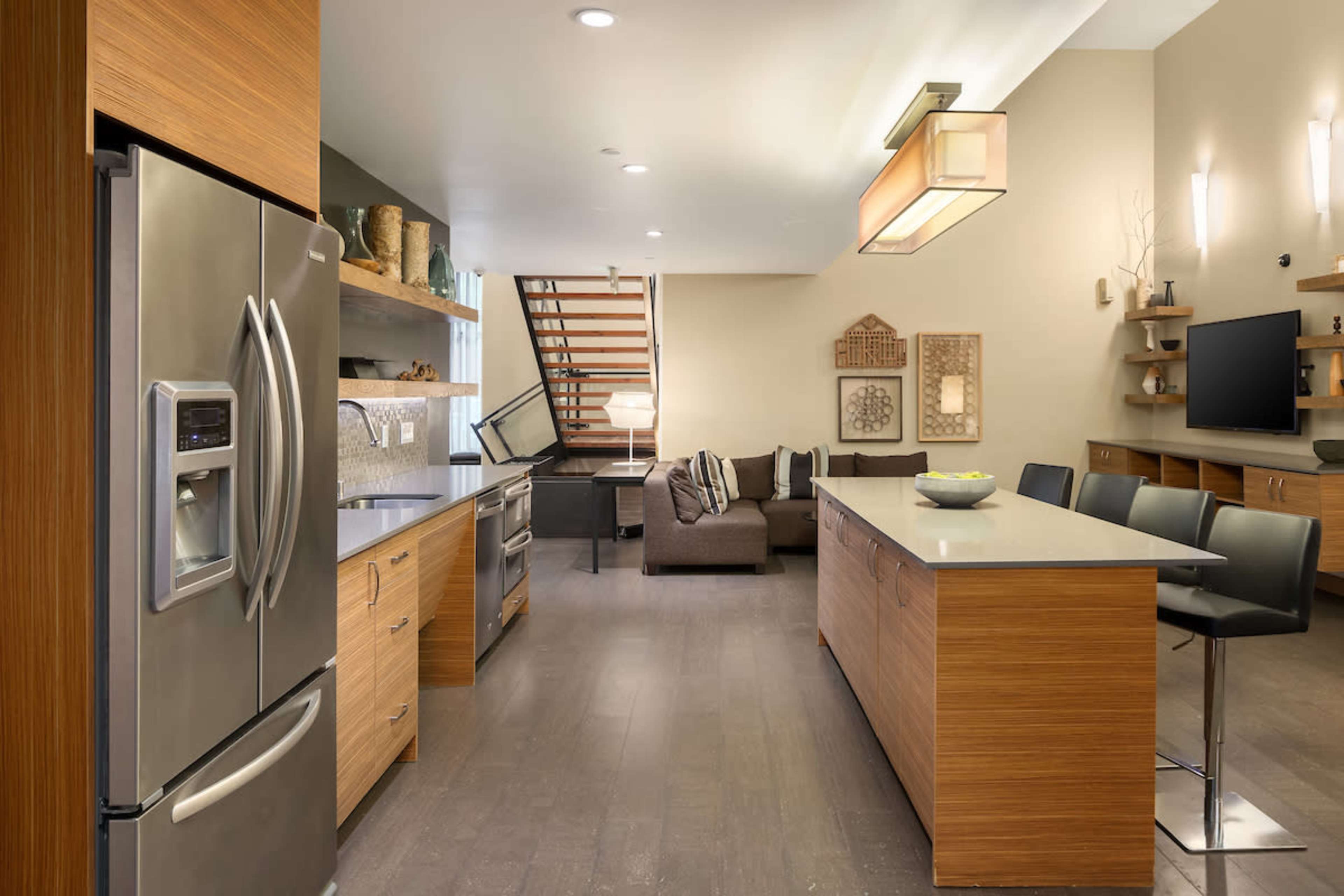 A modern kitchen and living area feature sleek stainless steel appliances, light wood cabinetry, an island with seating, and a staircase leading to an upper level.