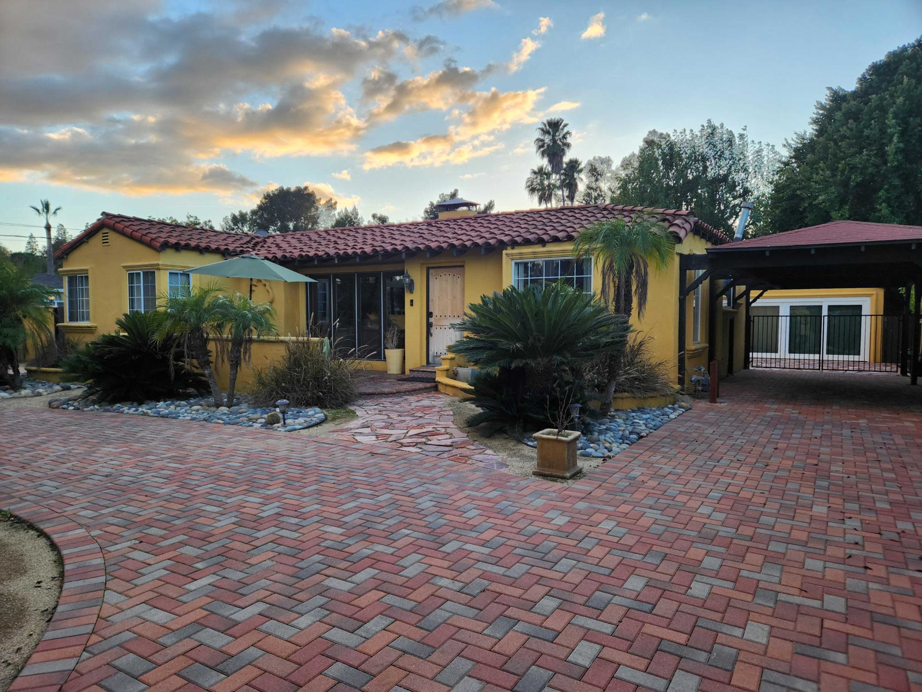 A yellow stucco house with a red tile roof is surrounded by palm trees and decorative plants, with a brick pathway leading to the entrance.