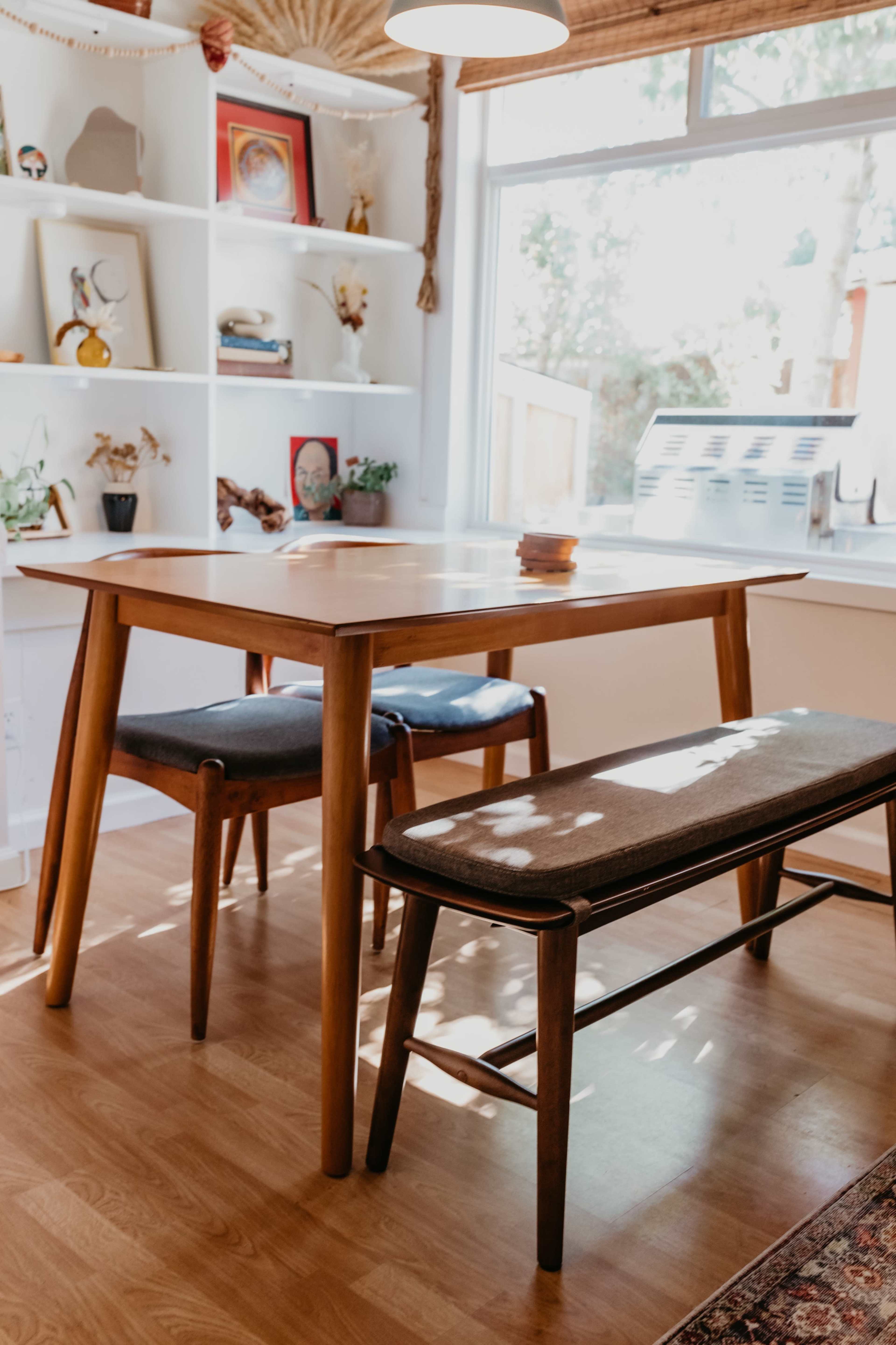 A wooden table with two chairs and a bench is positioned in a bright room filled with decorative items on shelves.