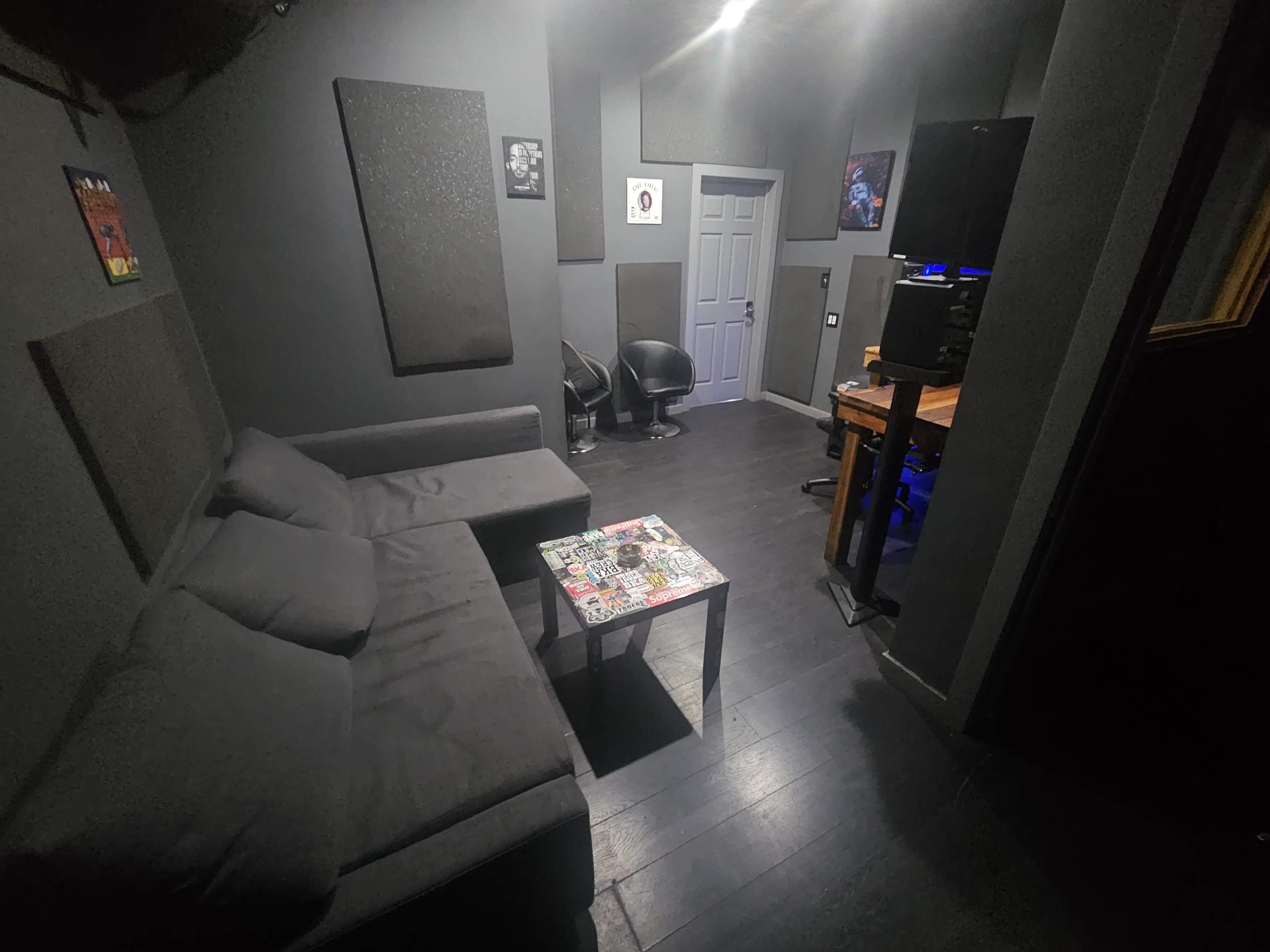 The image shows a dimly lit room with dark walls, featuring a gray sofa, a small table covered in stickers, and a workstation with a computer setup in one corner.