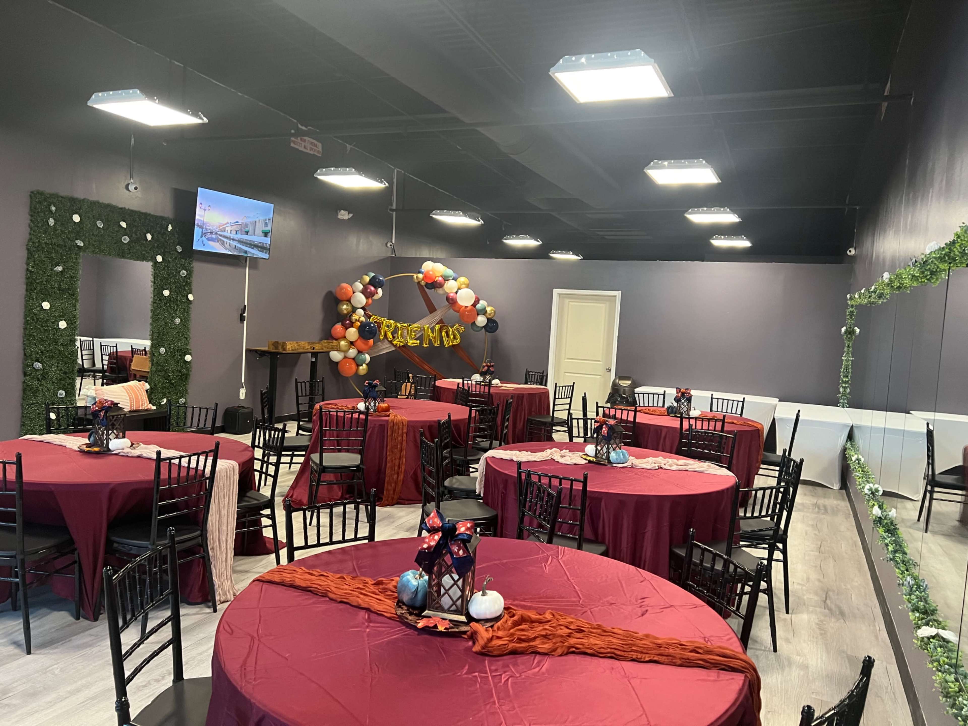 The image shows a banquet hall decorated with circular tables covered in red tablecloths, black chairs, and festive decorations, including balloons and a display table at the front.