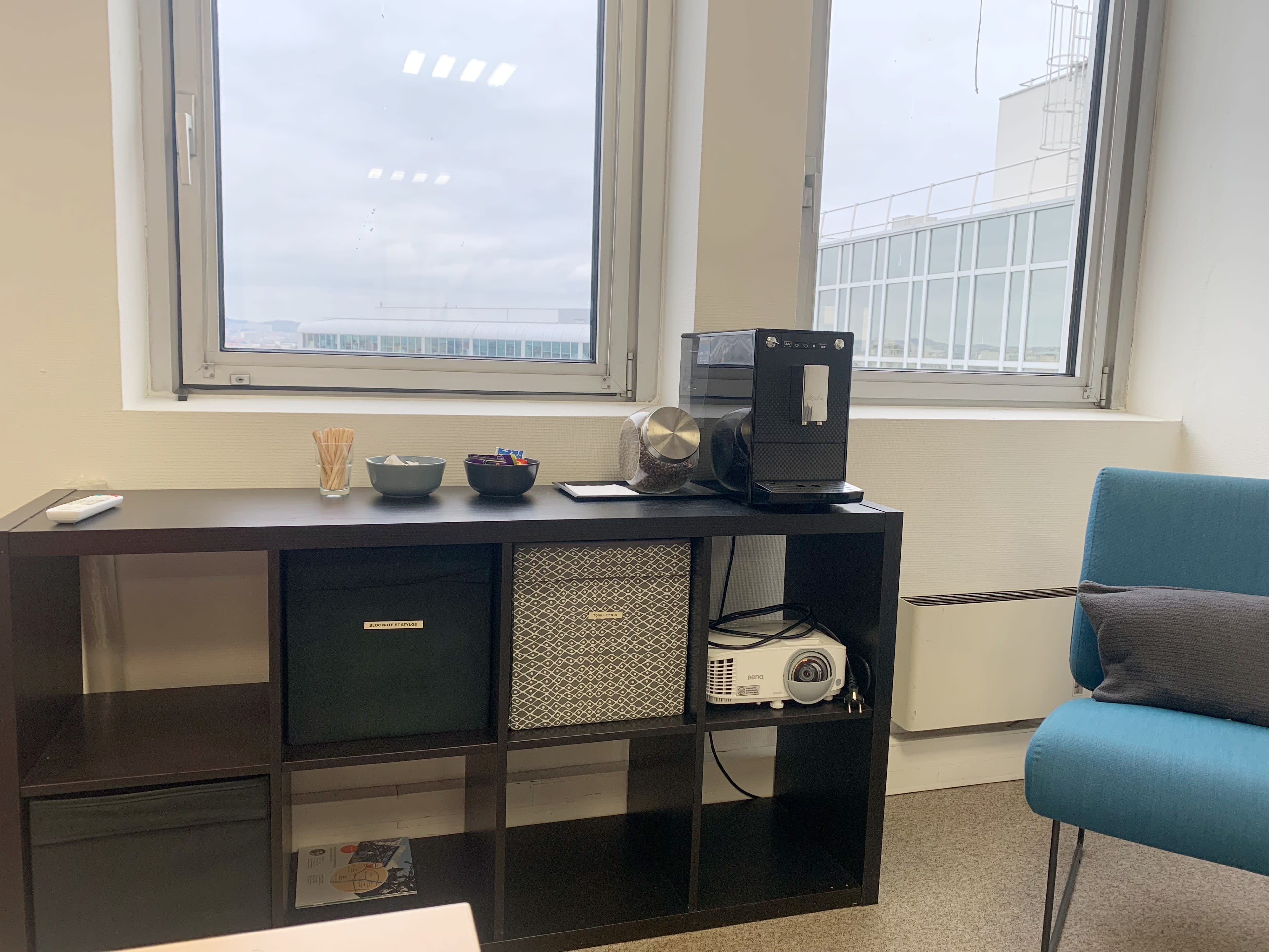 A workspace with a shelf holding a coffee machine, a projector, and storage boxes, alongside a blue chair and large windows overlooking an outdoor view.