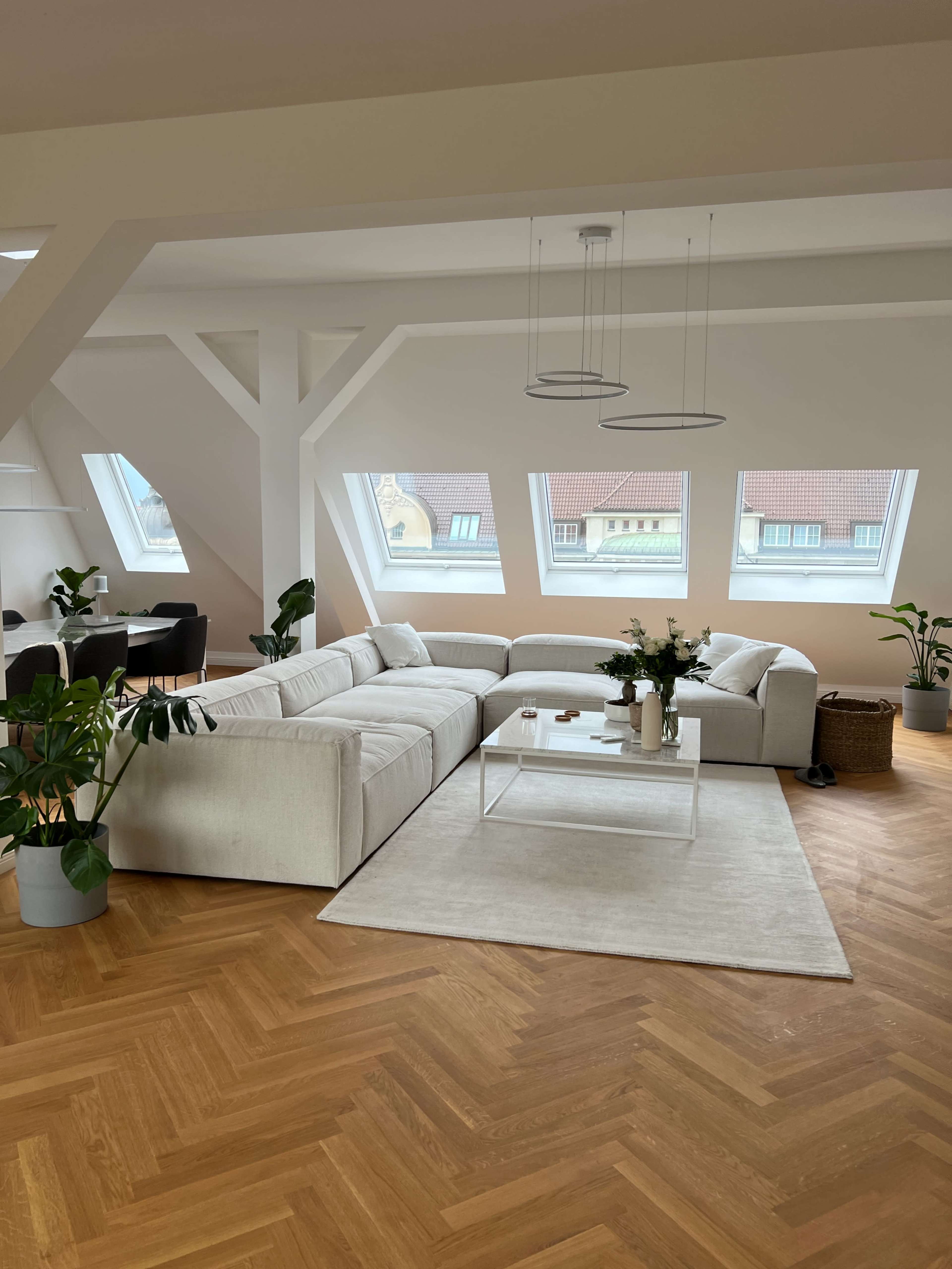 A spacious, modern living room with a large sectional sofa, a glass coffee table, and several plants, illuminated by natural light from multiple windows.