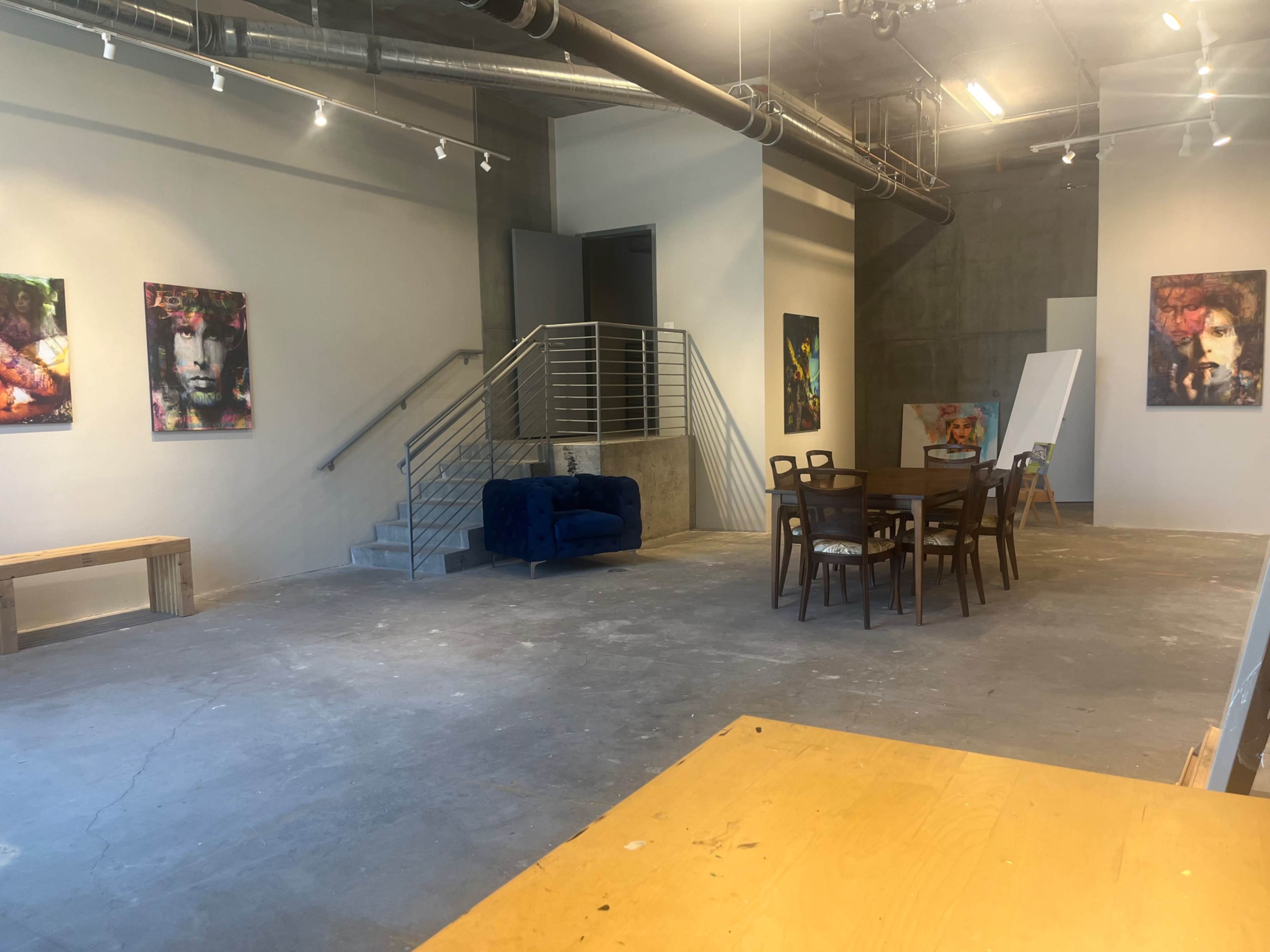 A spacious art studio with a table and chairs, a blue couch, and several colorful paintings on the walls.