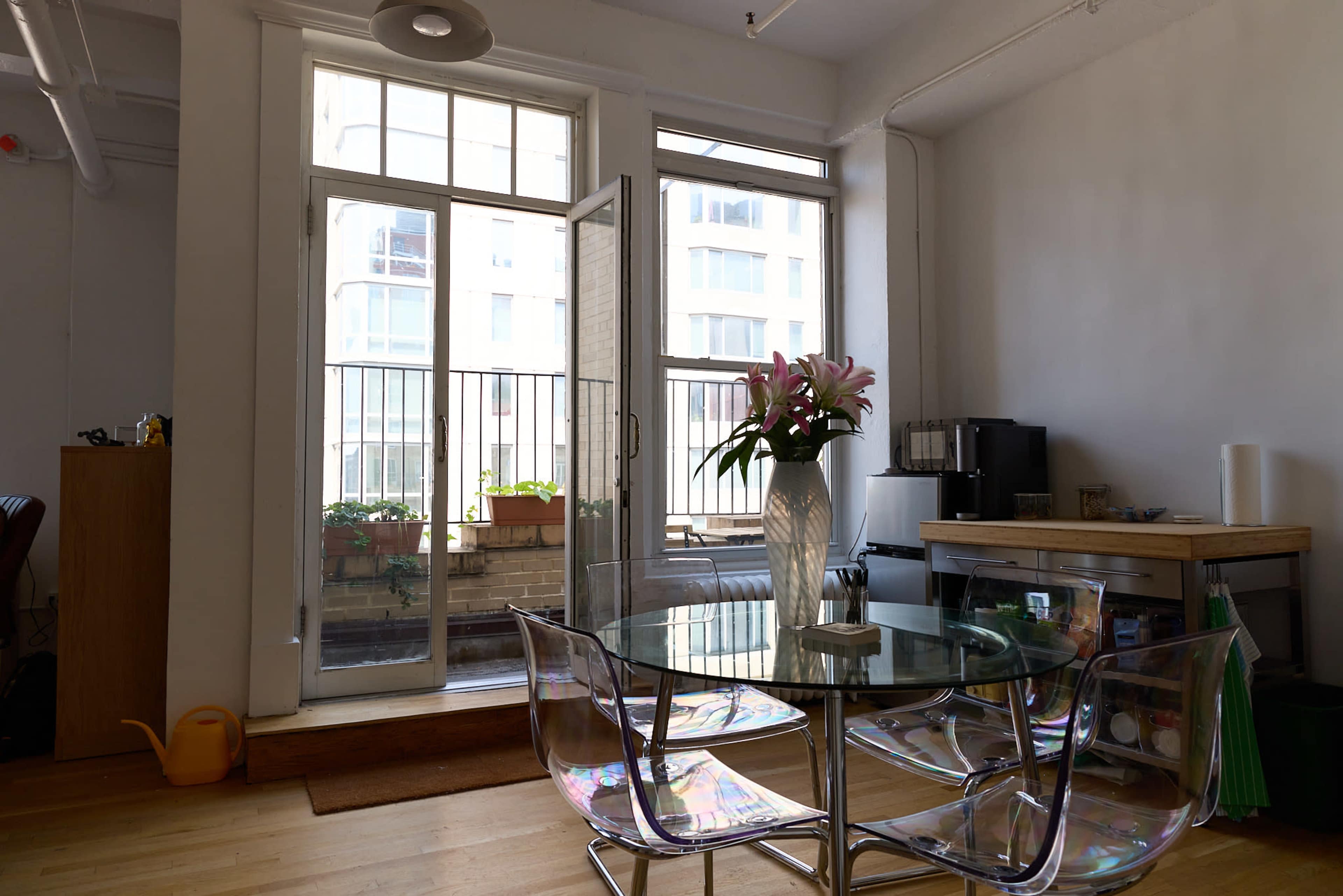 A clear glass dining table with plastic chairs is positioned in an apartment featuring large windows that open to a balcony filled with potted plants.