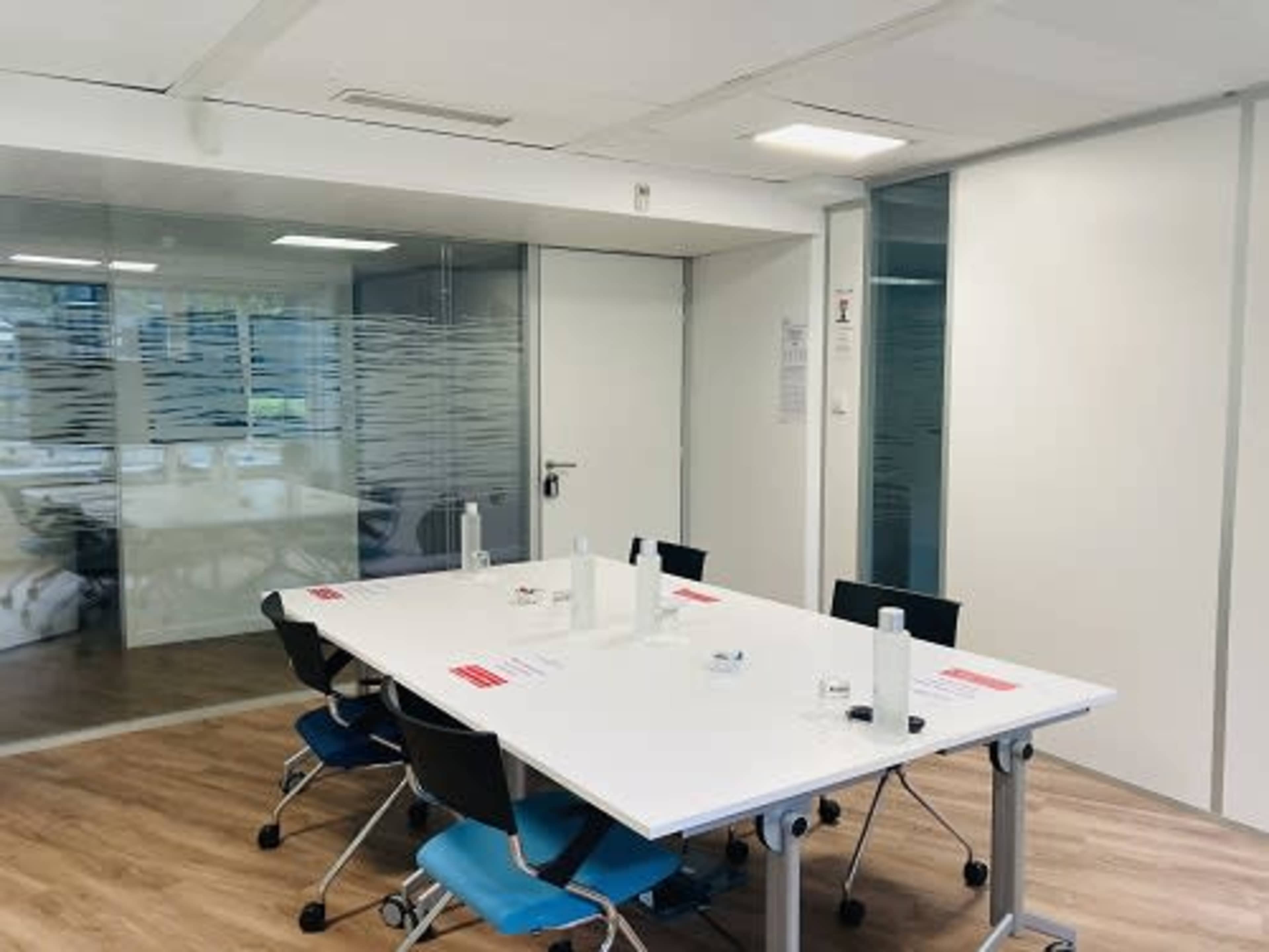 A modern meeting room features a large white table surrounded by black and blue chairs, with glass walls and a view of a window in the background.