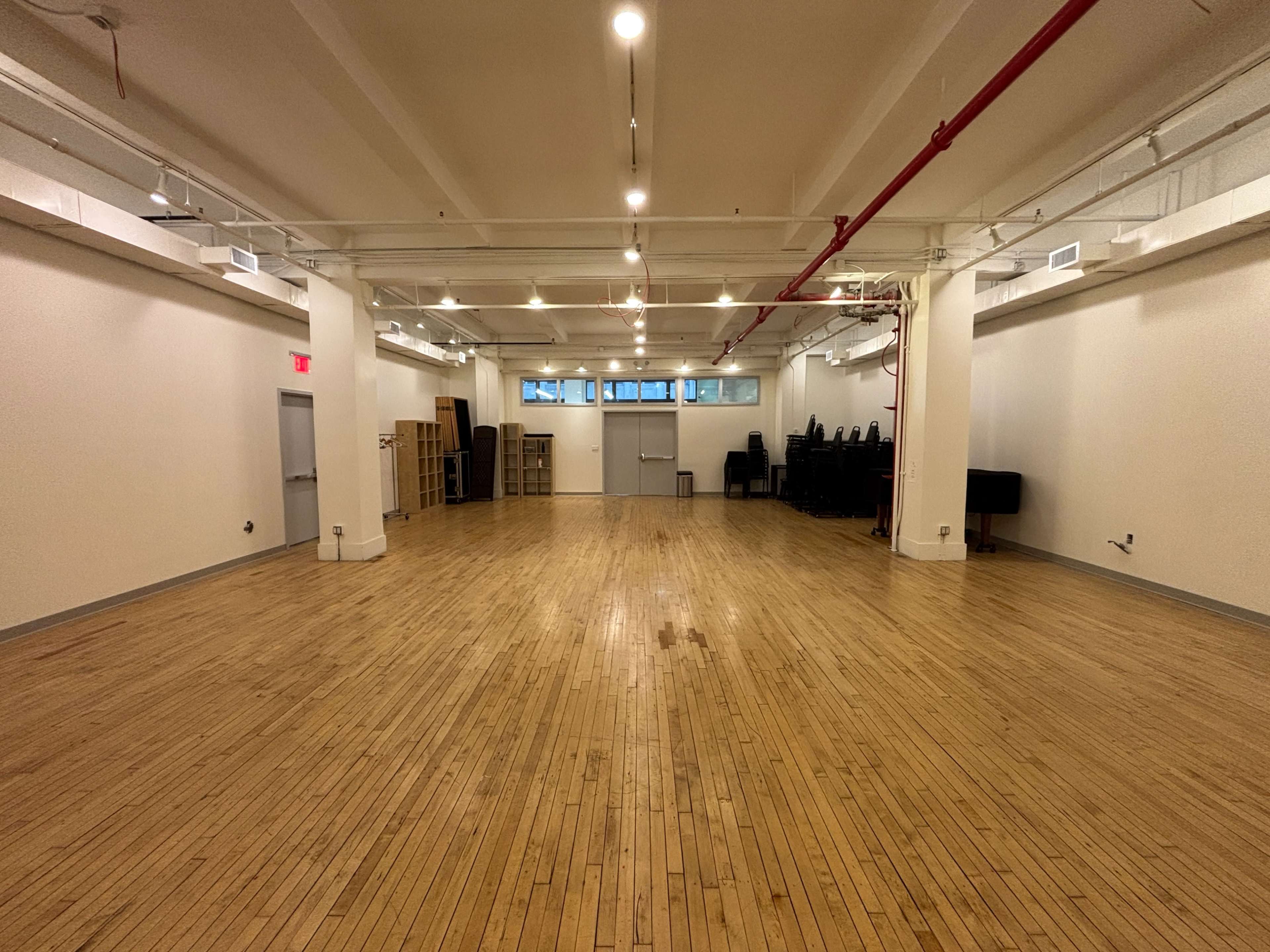 The image shows a spacious, empty room with polished wooden flooring and large windows, featuring stacked chairs in the back corner.