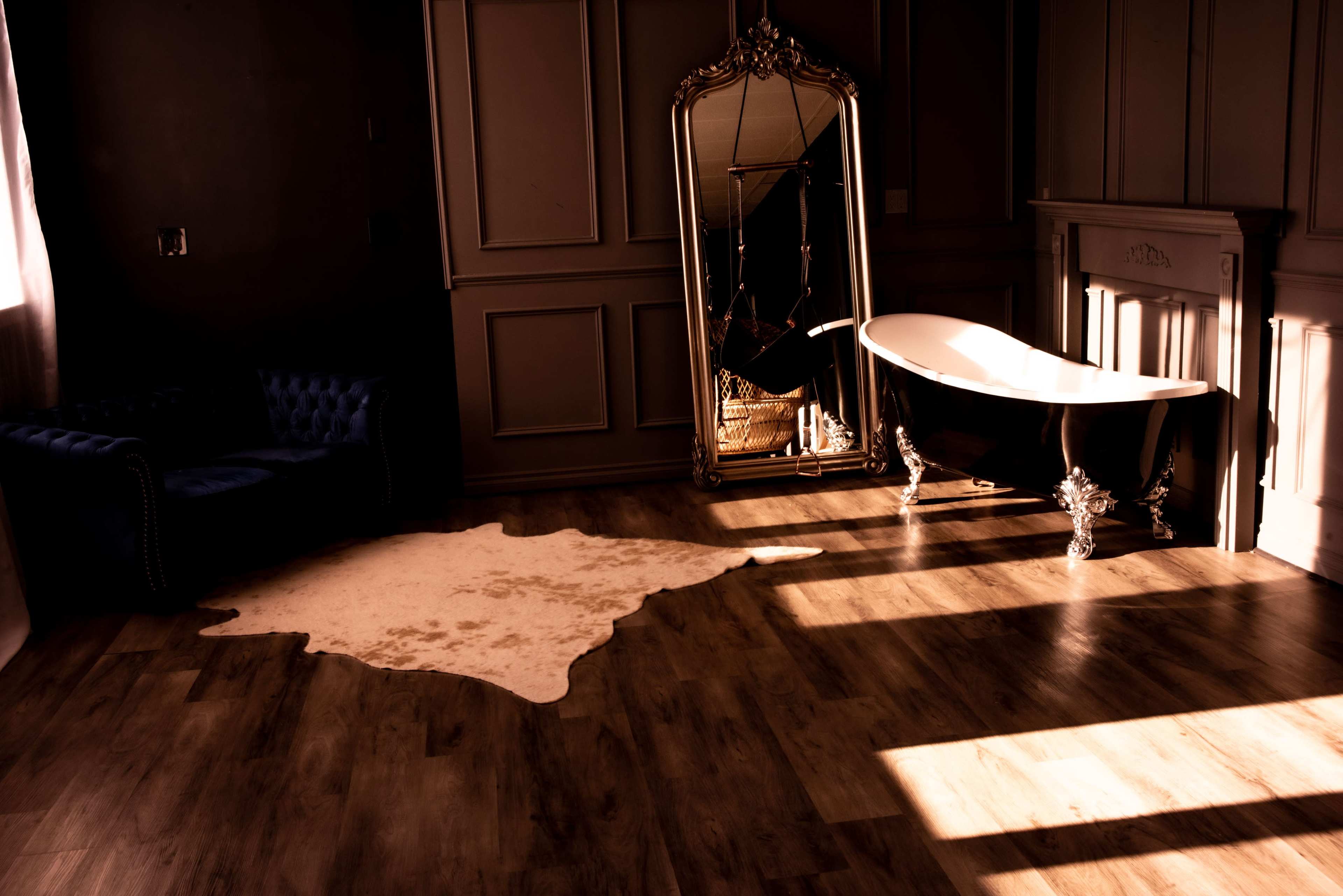 A stylish bathroom featuring a freestanding black and white bathtub, a large ornate mirror, a plush blue sofa, and a light-colored area rug on wooden flooring.