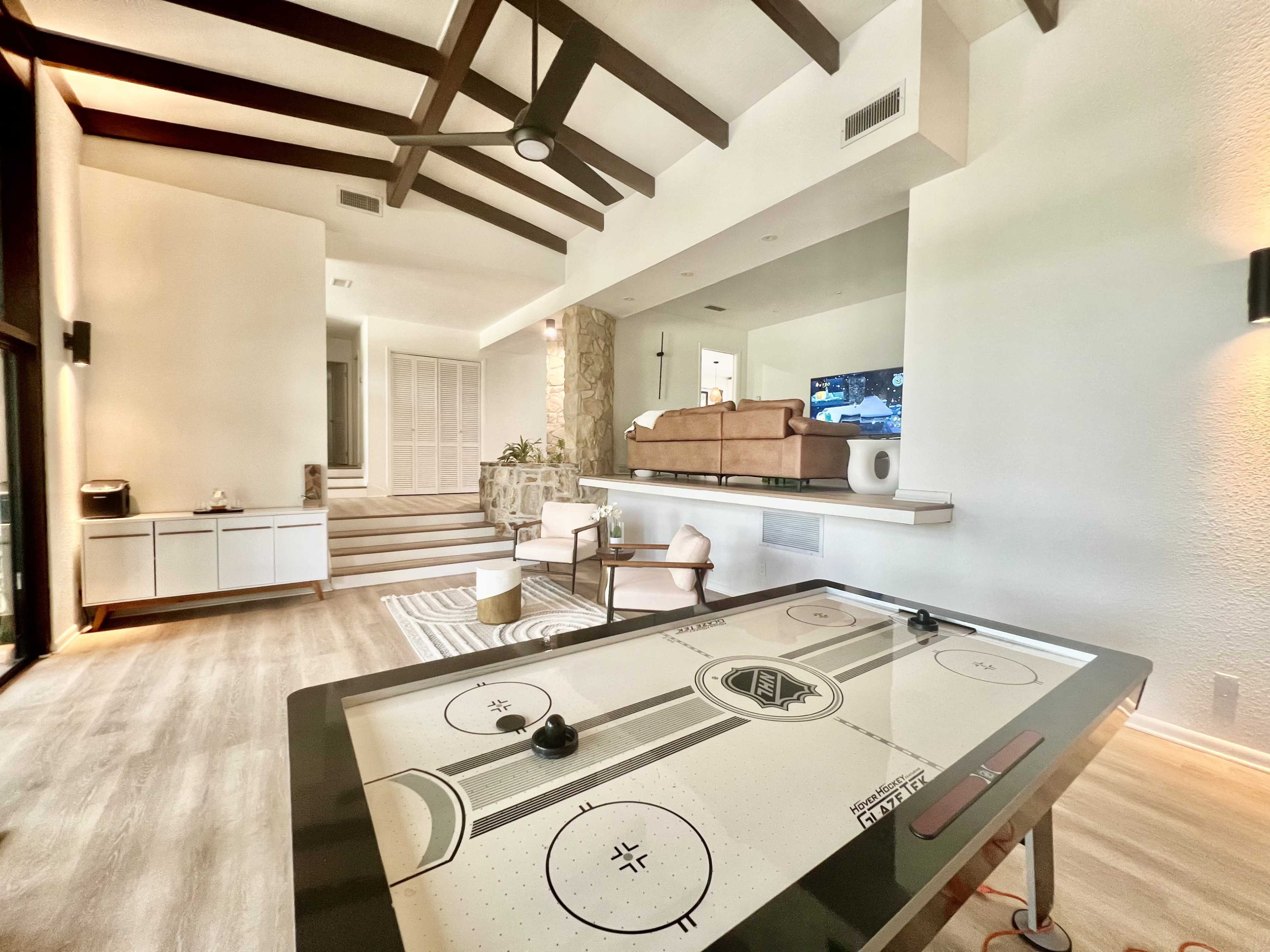 A modern living area featuring an air hockey table, a stone fireplace, and brown furniture against a backdrop of exposed wooden beams.
