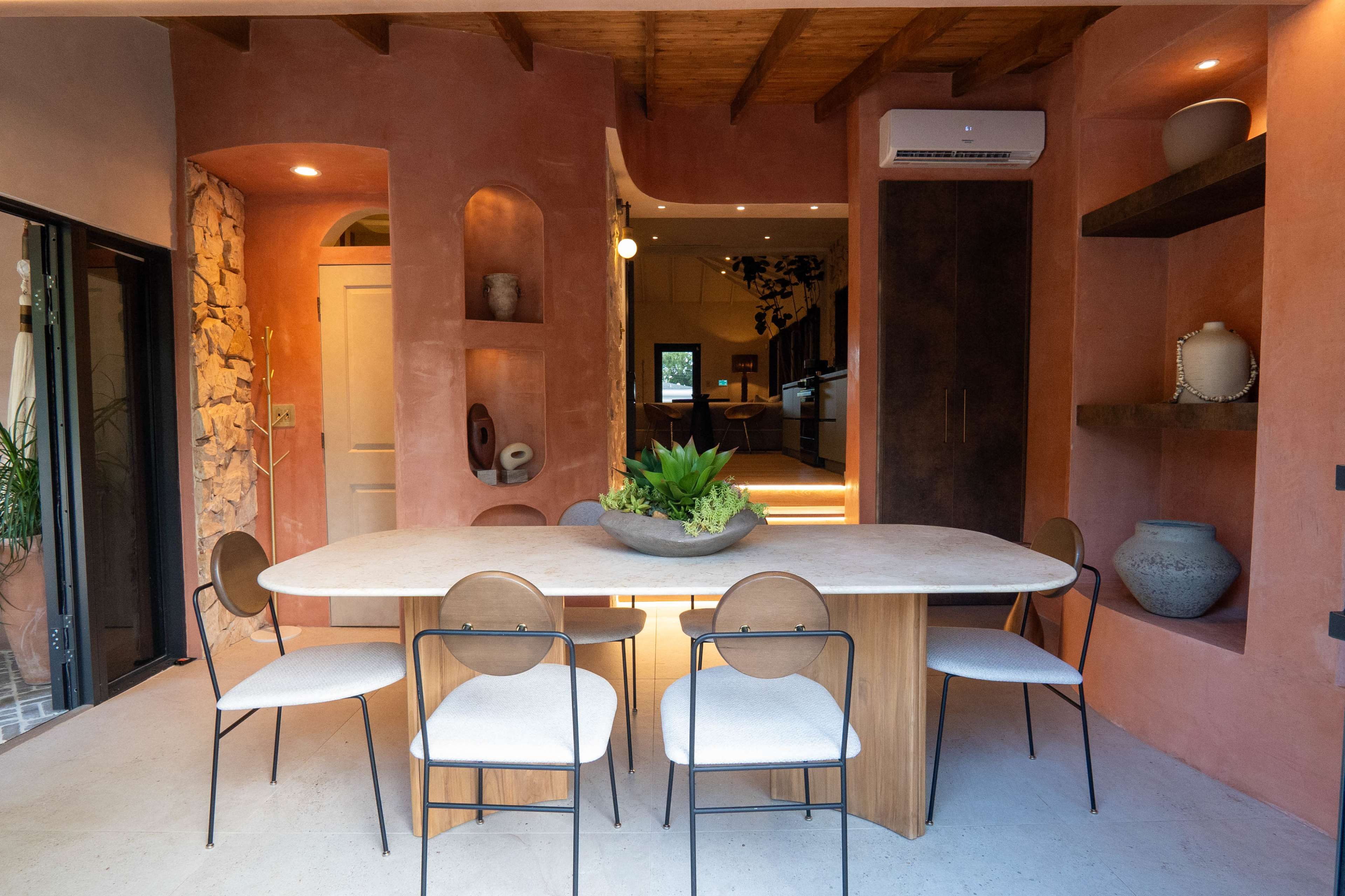 The image shows a modern dining area with a large wooden table surrounded by six chairs, and walls featuring a warm, pink-toned plaster finish.