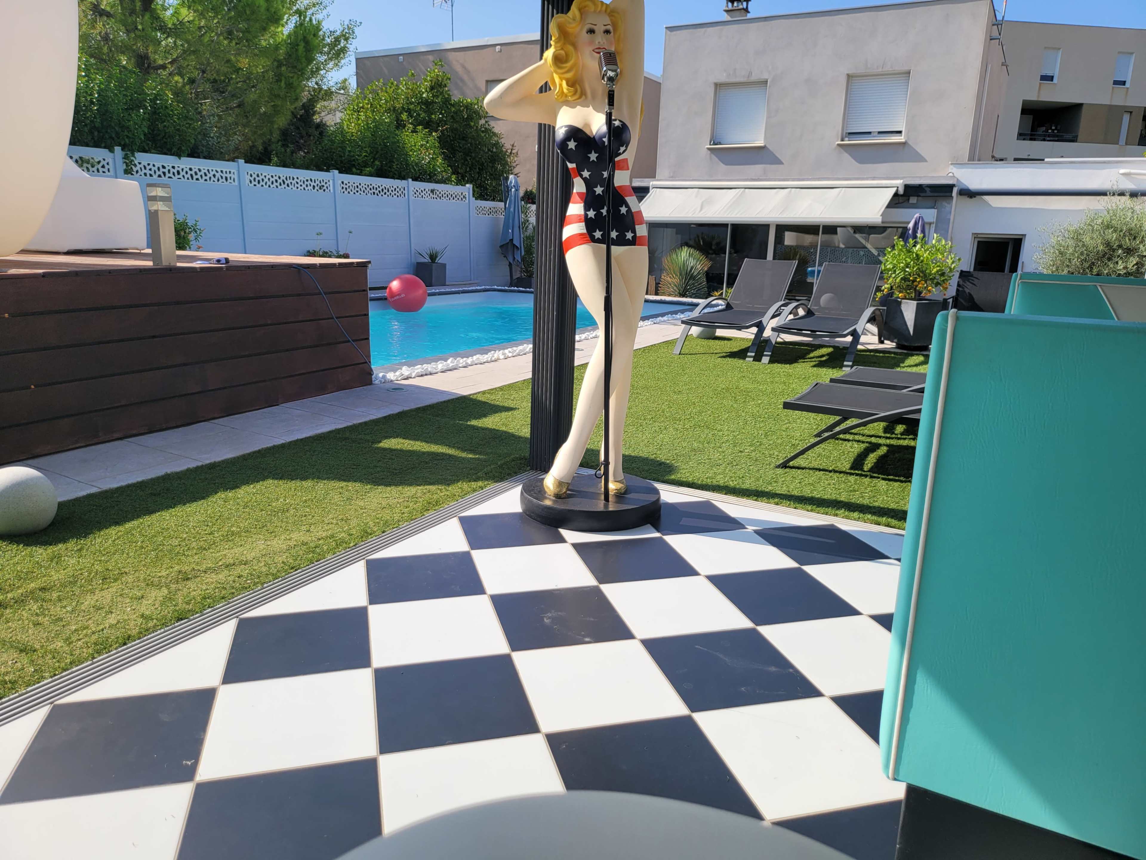 A pool area features a checkered floor and a vintage mannequin in a striped swimsuit, with lounge chairs and a blue pool in the background.