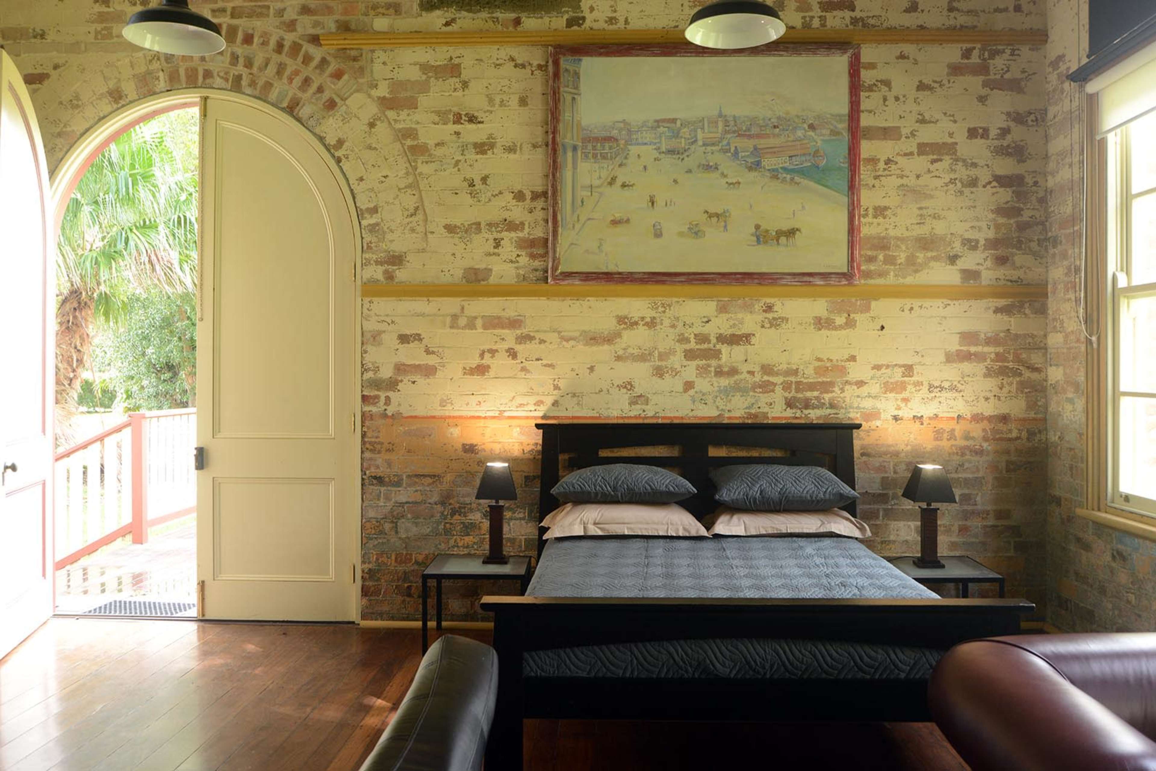 The room features a bed with blue and white bedding, flanked by two lamps, and a large painting above it, with an arched doorway leading outside.