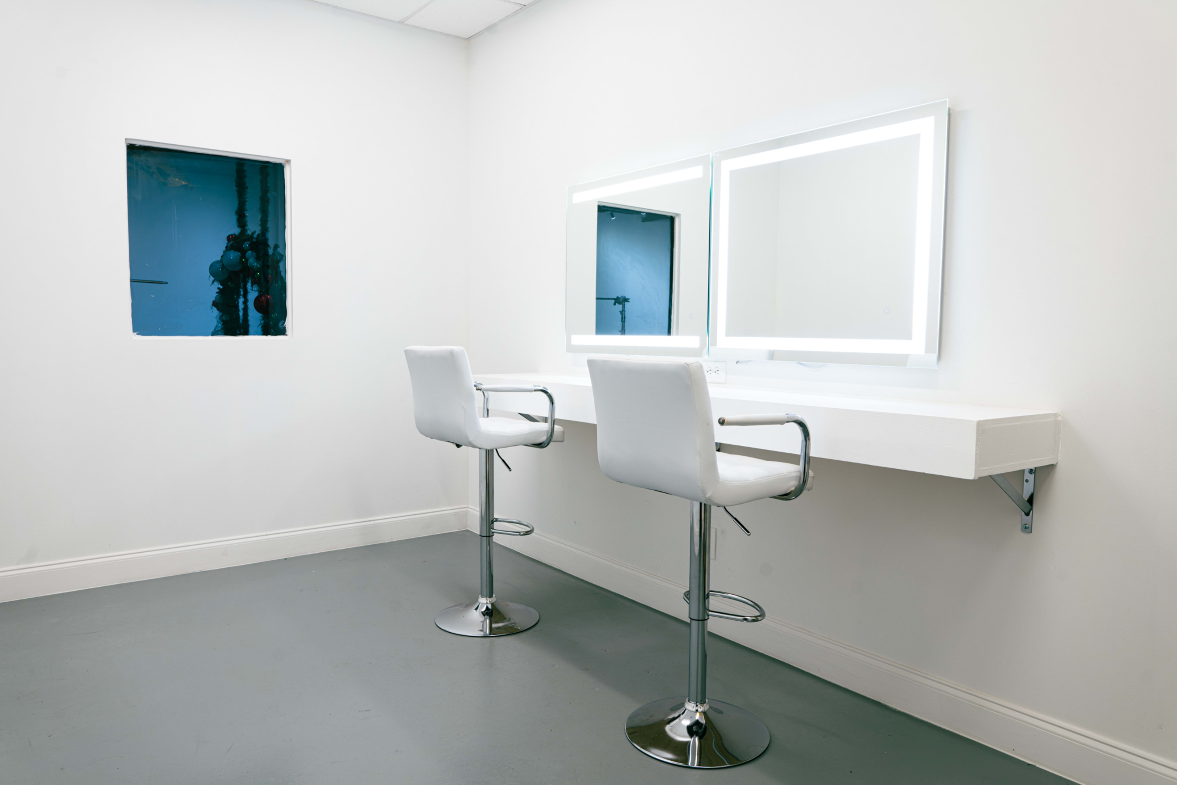 The image shows a minimalist makeup station with two white bar stools and backlit mirrors in a neutral-toned room.