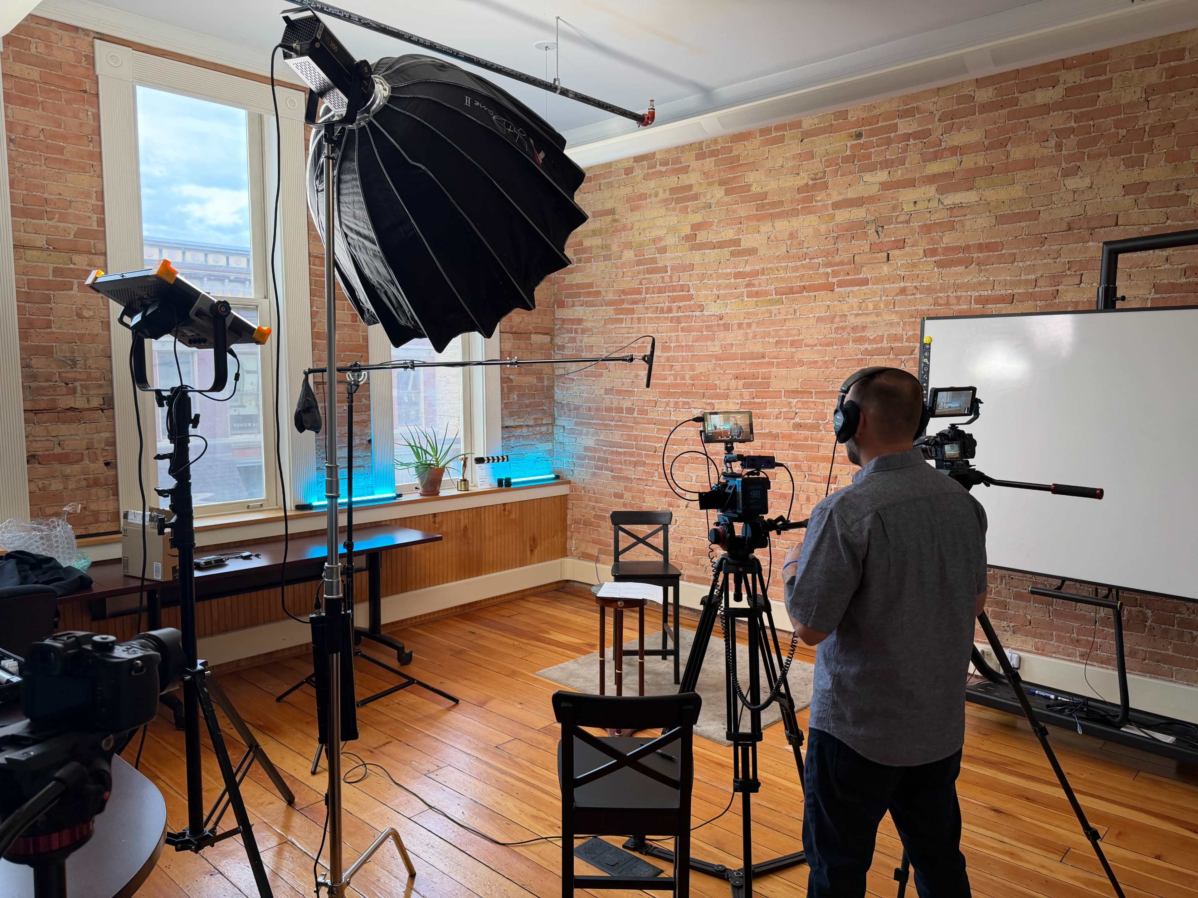 A man stands behind multiple cameras in a room with brick walls, preparing for a video shoot with lighting equipment and a whiteboard in the background.