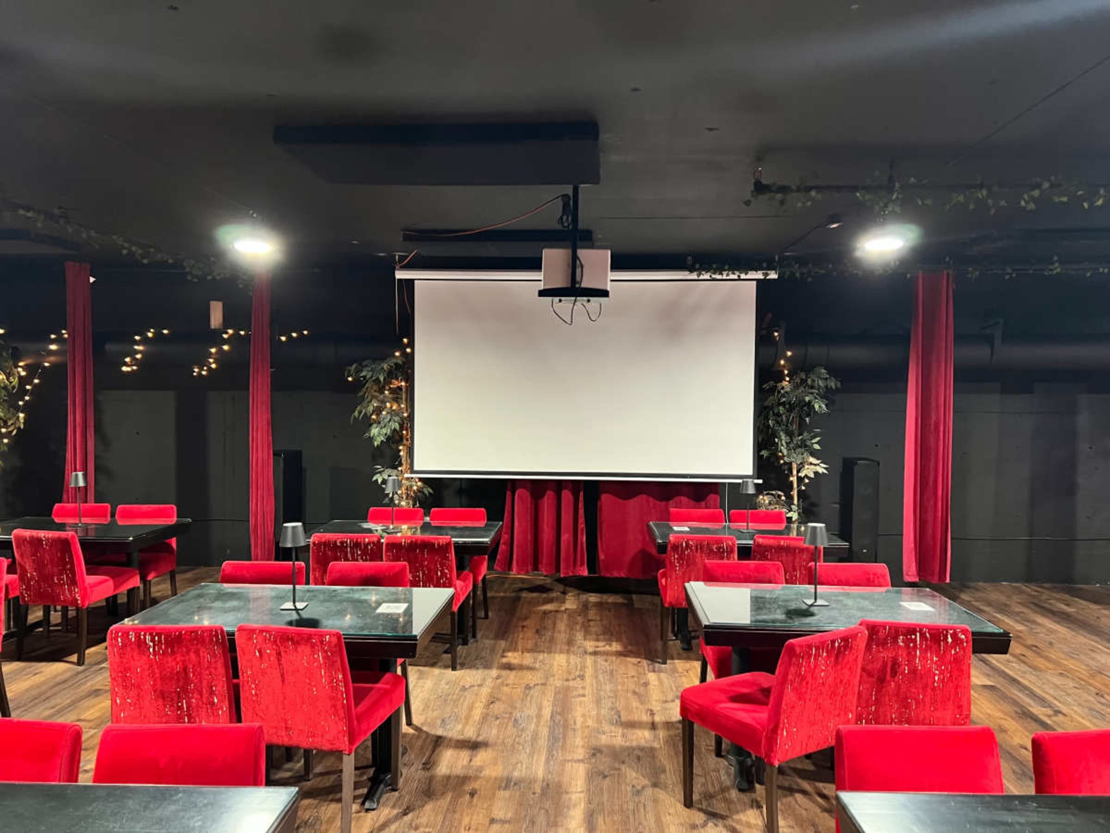 The image shows a dimly lit venue with red velvet seating, wooden floors, and a projector screen at the front.