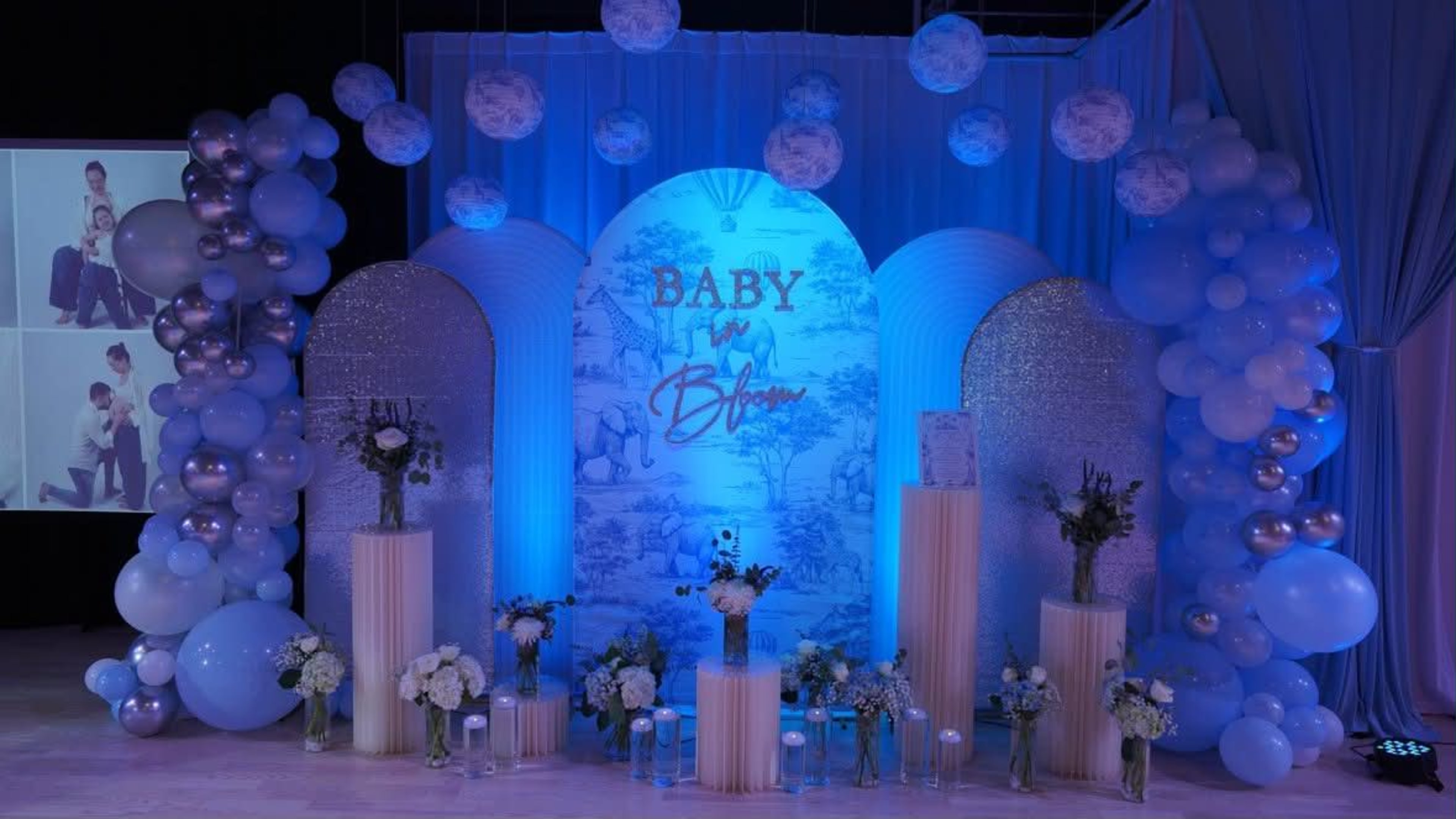 The image features a decorated stage for a baby shower, complete with blue and white balloons, floral arrangements, and a backdrop displaying the words "Baby is Brewing."