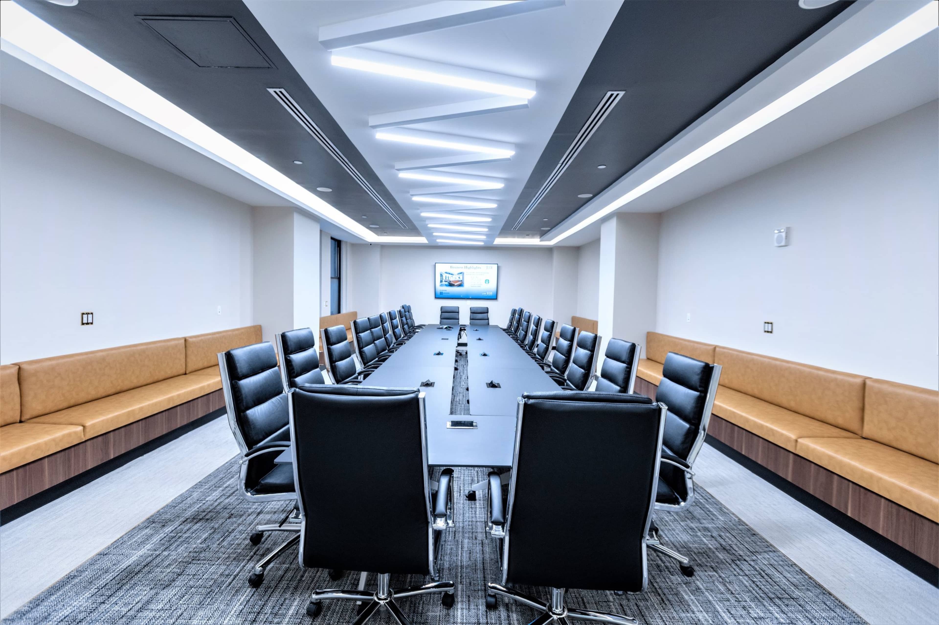 A modern conference room features a long table surrounded by ergonomic black chairs and a presentation screen at one end, illuminated by linear ceiling lights.