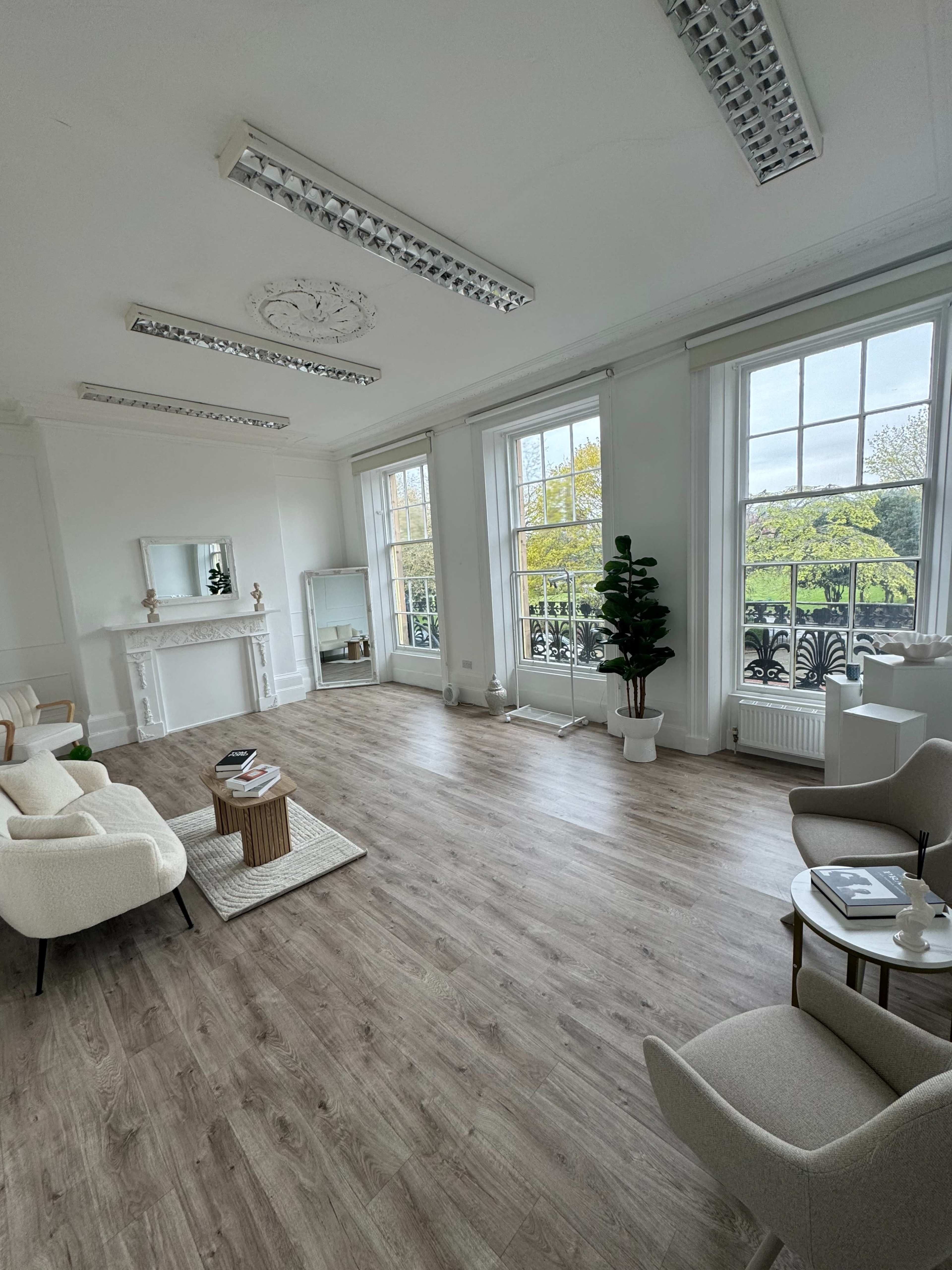 The image shows a spacious, well-lit room with large windows, light wood flooring, and minimalist furniture, including two chairs and a coffee table.