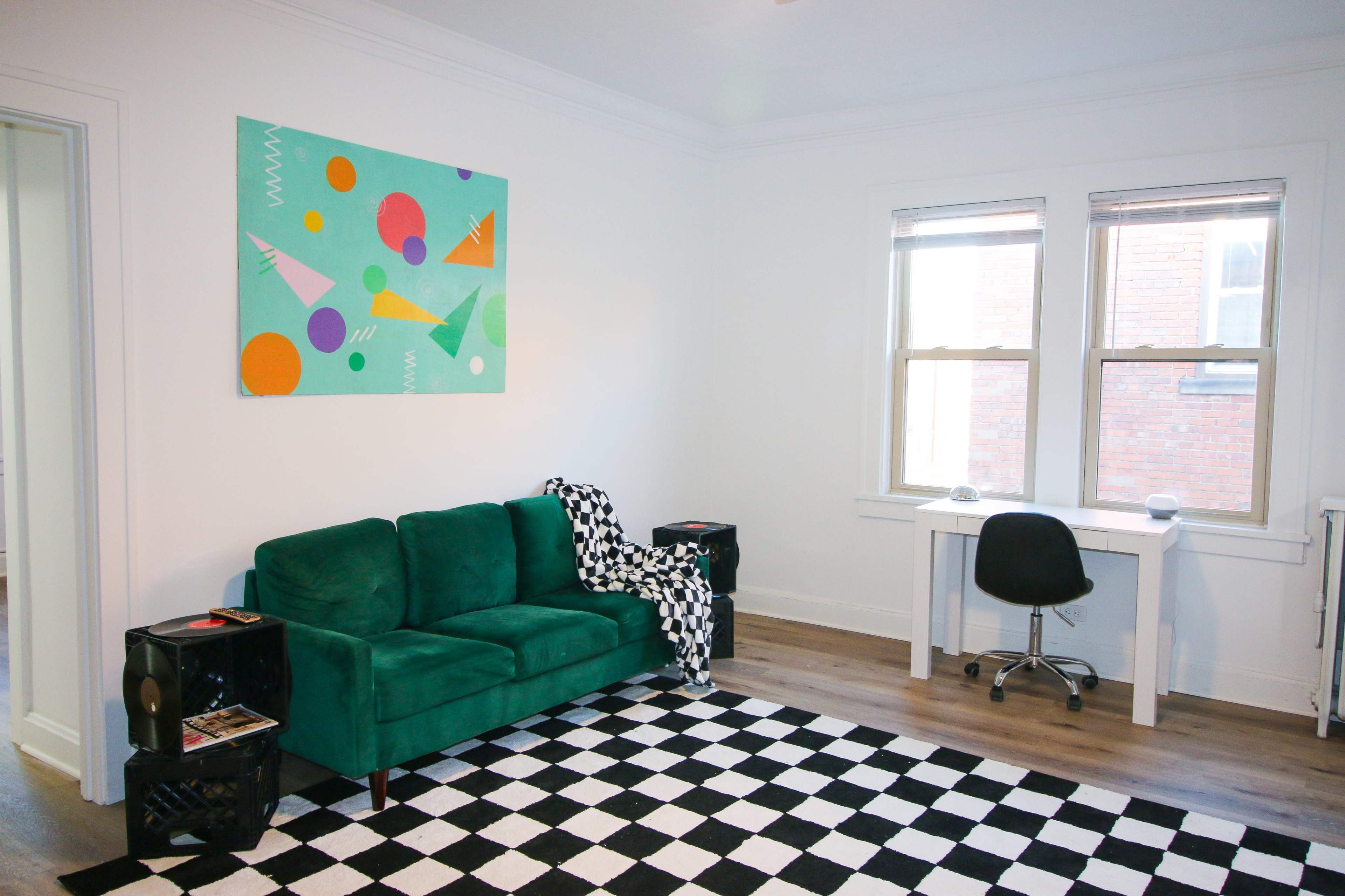 The room features a green sofa, a black and white checkered rug, a white desk with a chair, and an abstract painting on the wall.