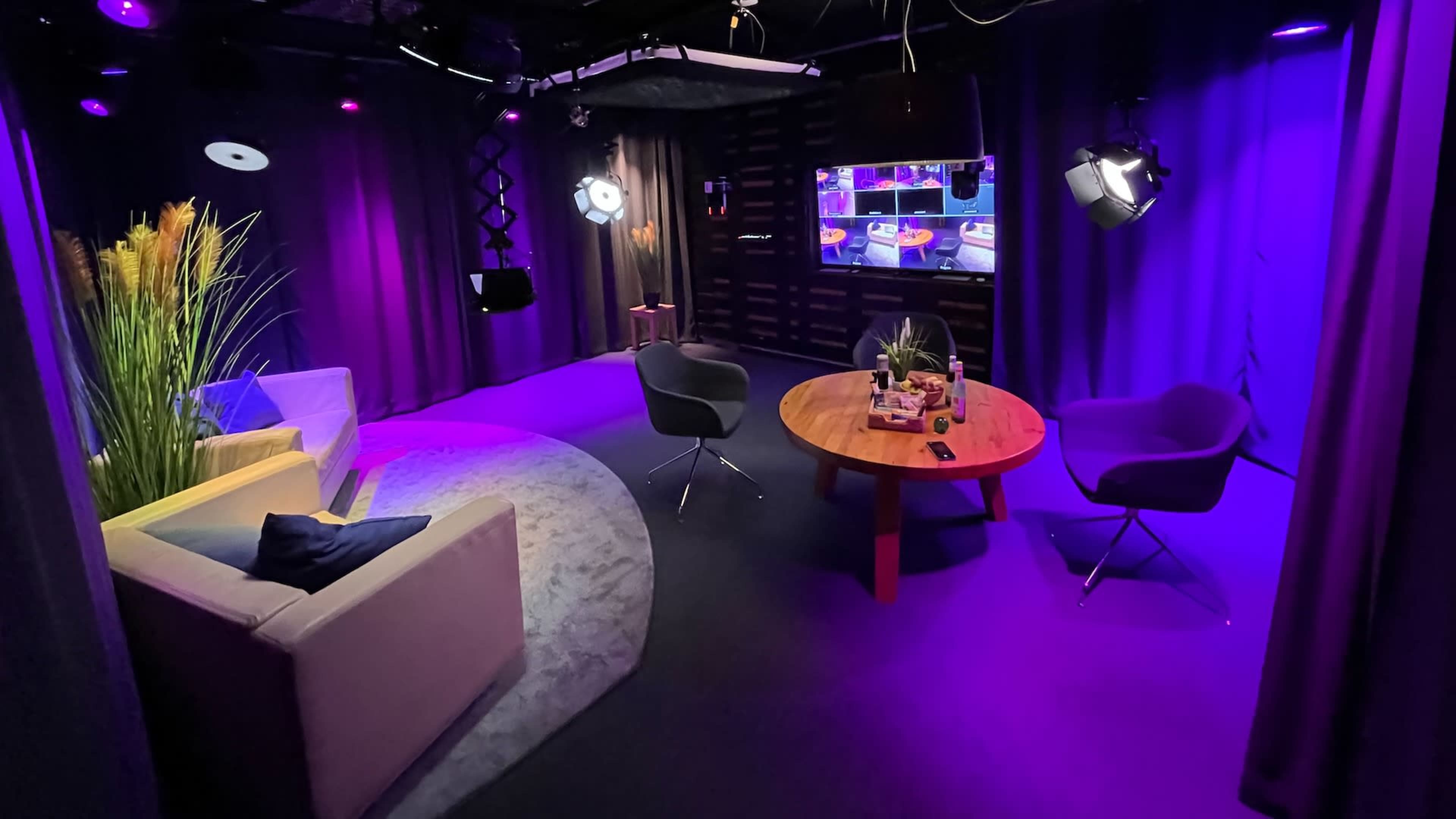 The image shows a television studio with a circular area rug, a sofa, two chairs, a wooden table, and multiple lighting fixtures, all set against a backdrop of dark curtains.