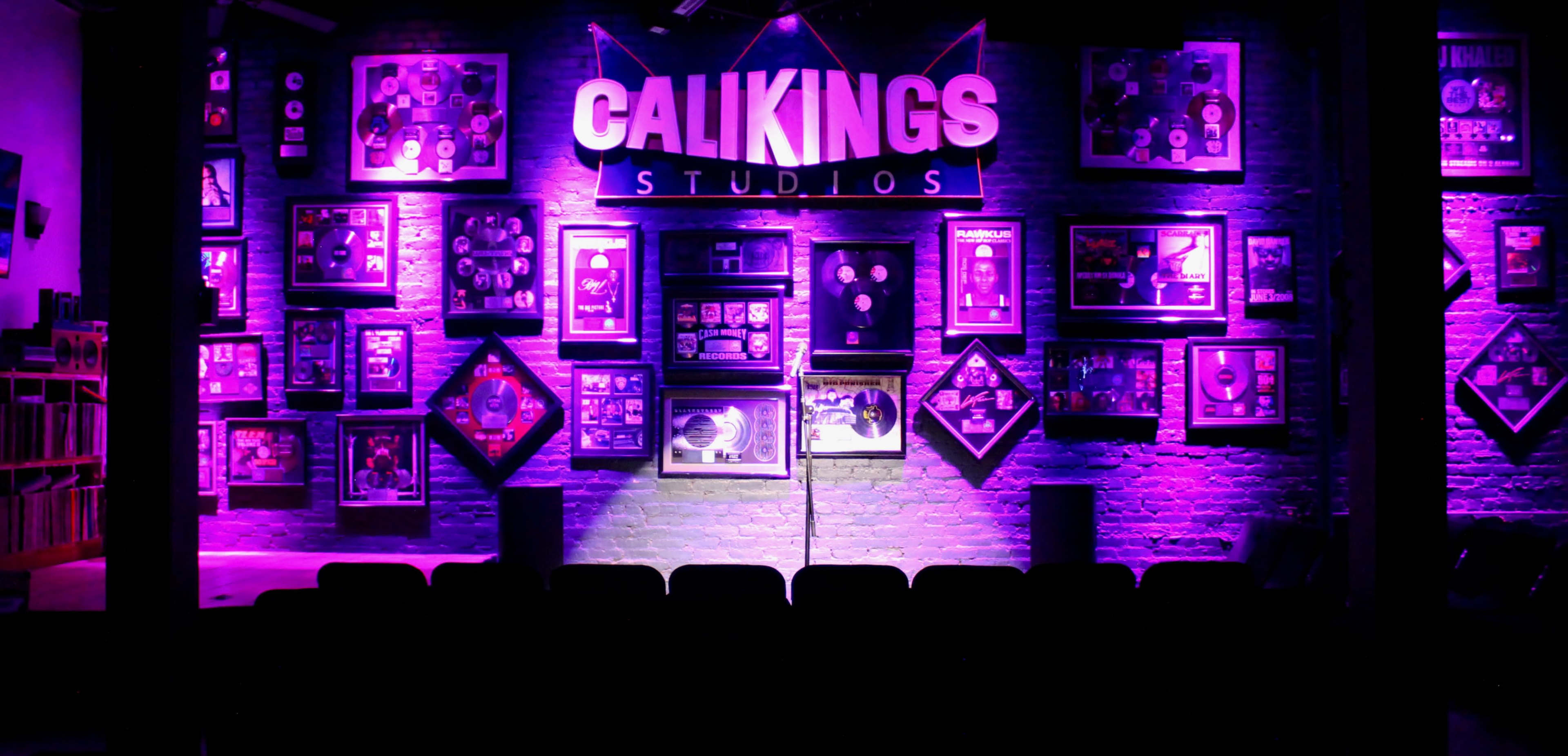 A performance stage with a microphone in front of a brightly lit wall displaying framed records and awards at Calikings Studios.