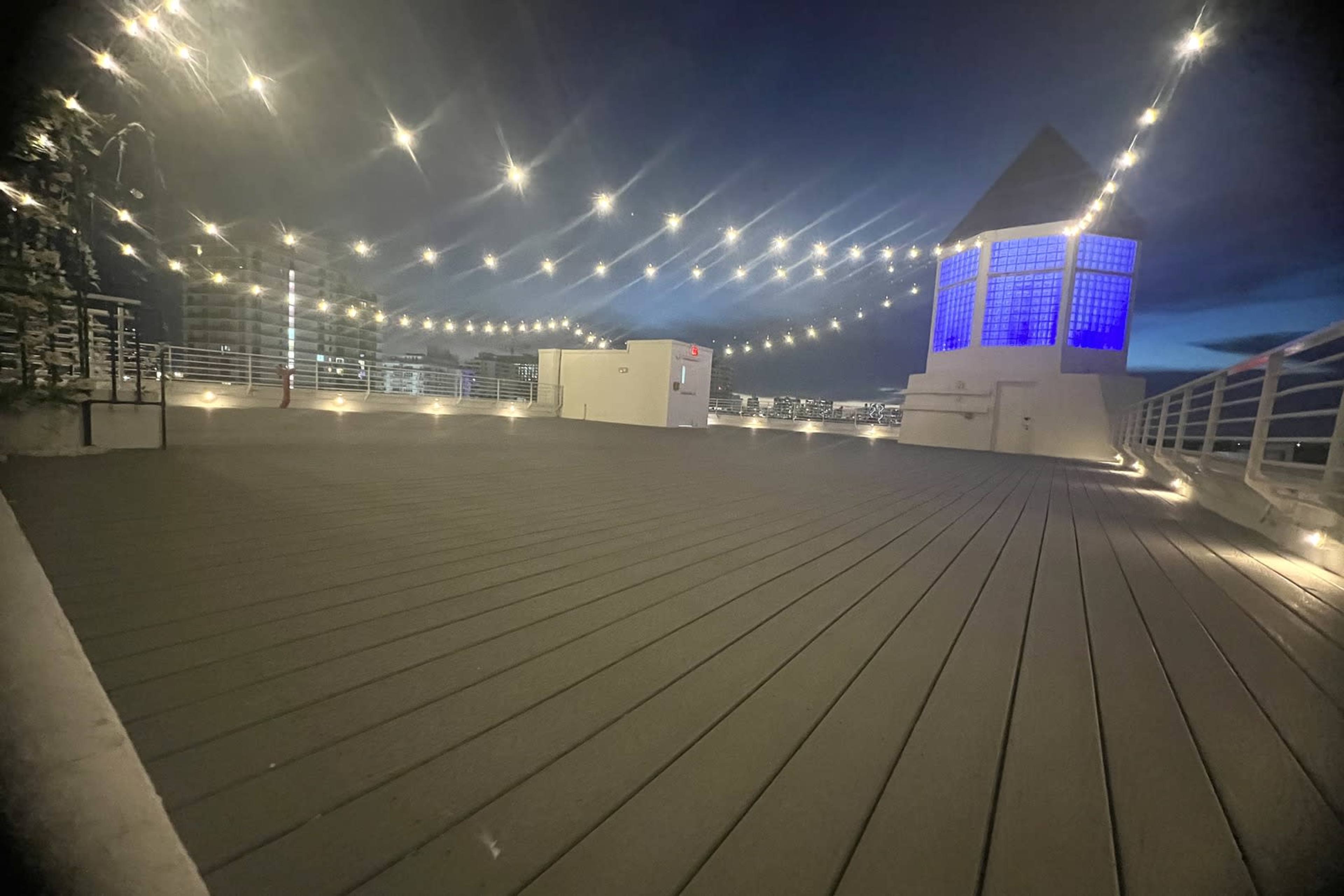 The image shows a rooftop area adorned with string lights and a structure featuring a blue-lit tower under a twilight sky.