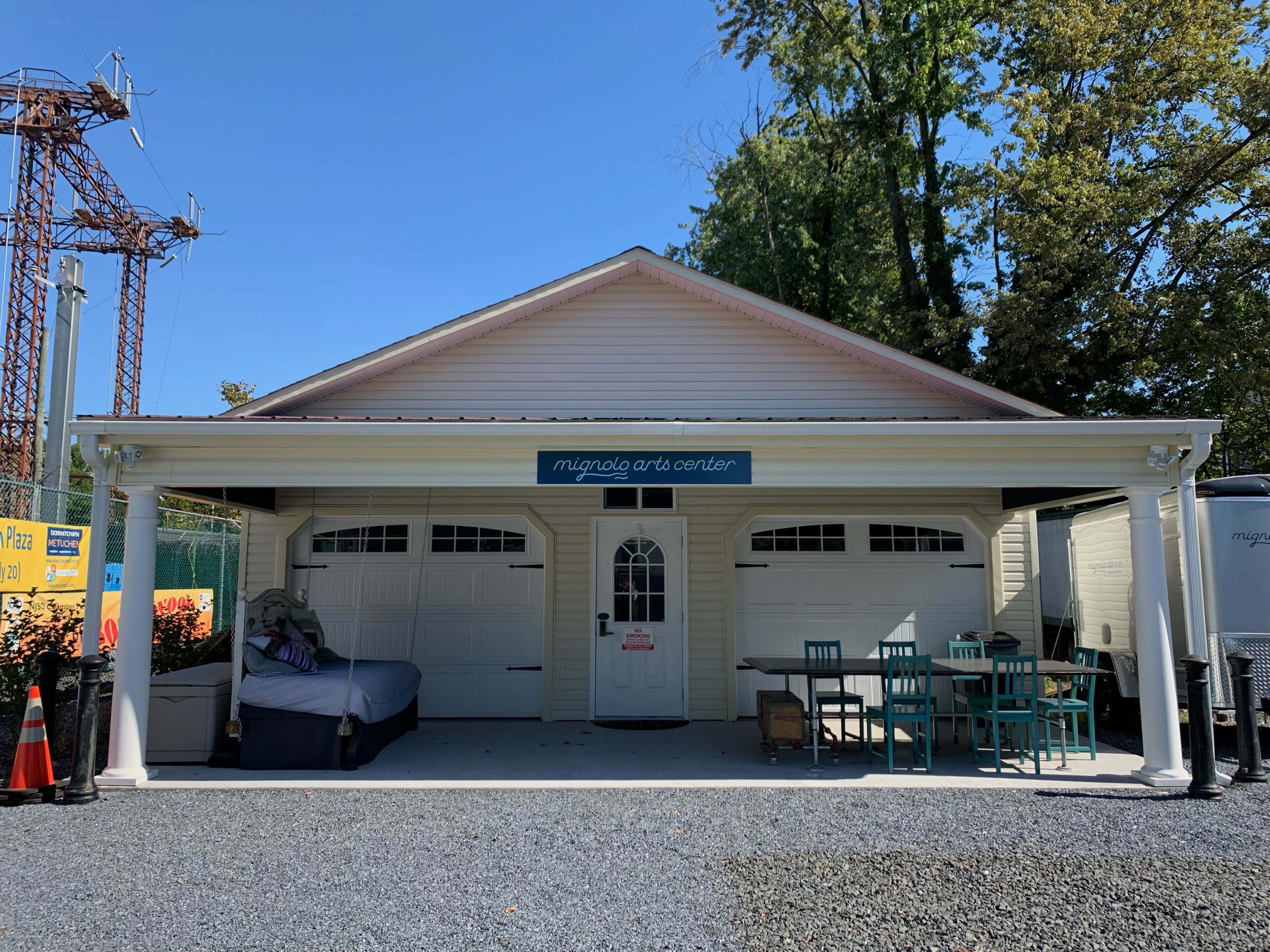 The image shows a building with a sign reading "mugnato arts center," featuring two garage doors and a table with chairs outside on a gravel surface.